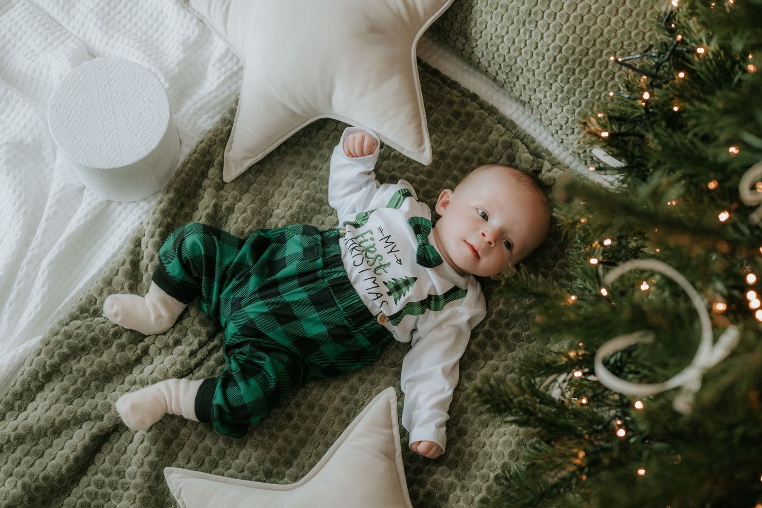 Baby in green plaid Christmas outfit lying on a green blanket with star pillows next to a decorated Christmas tree.
