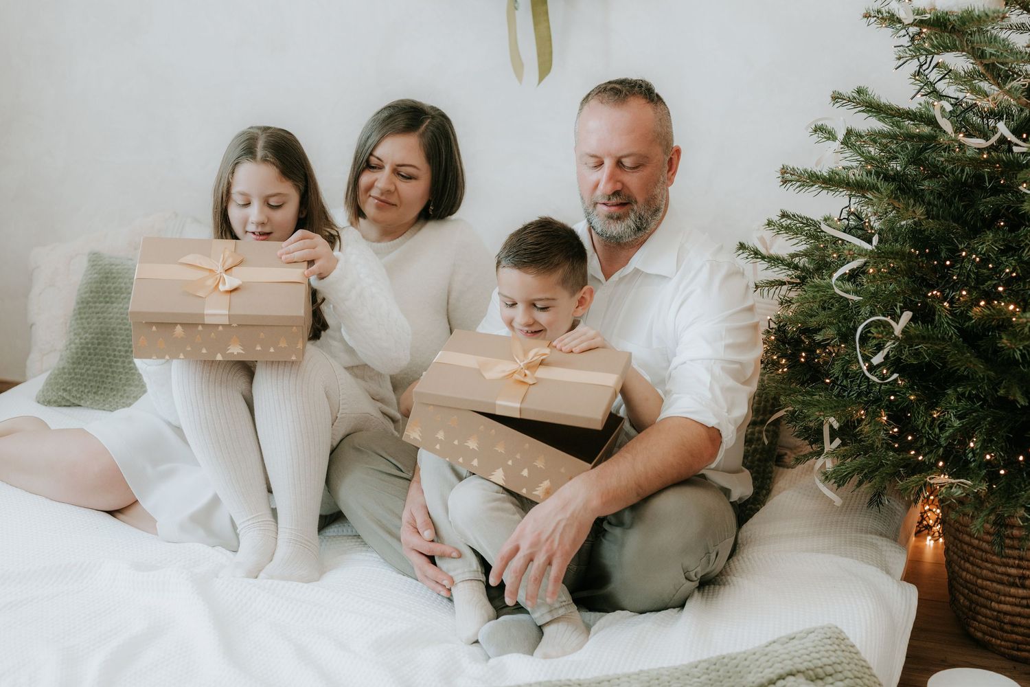 Family of four exchanging Christmas gifts near decorated tree in cozy white holiday setting.