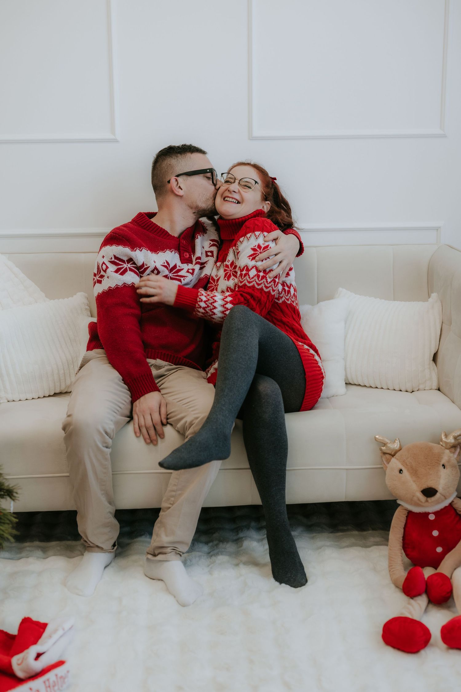 Couple in matching red Nordic sweaters sharing a kiss on a white sofa with Christmas decorations nearby.