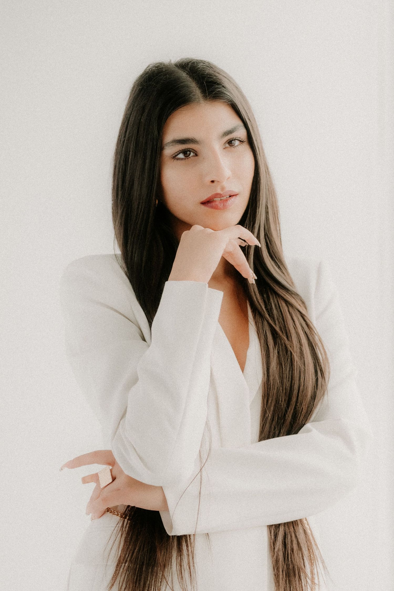 Young woman with long dark hair in white blazer posing thoughtfully against white background.