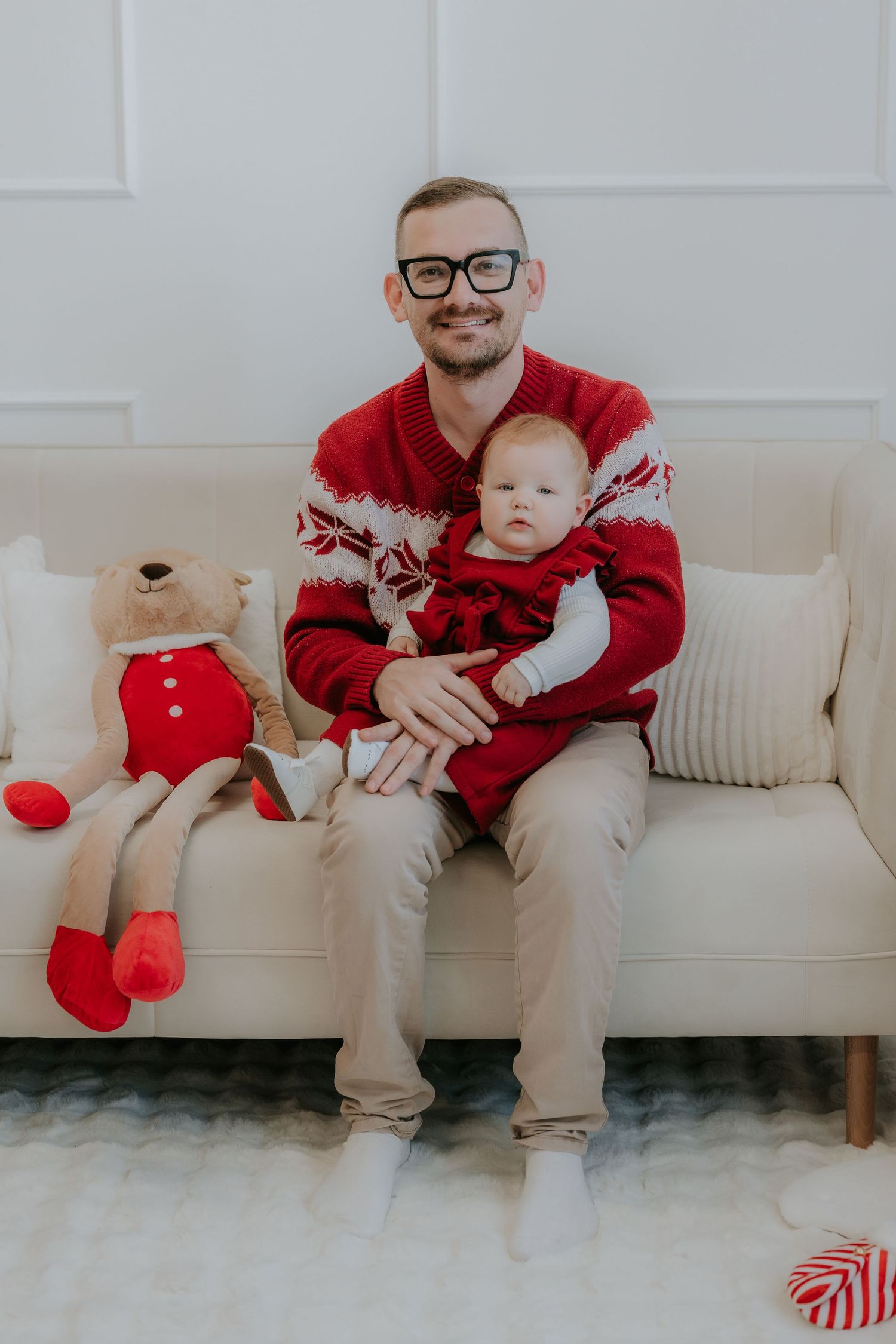 Man in red holiday sweater holding baby on cream sofa beside teddy bear in Christmas pajamas.