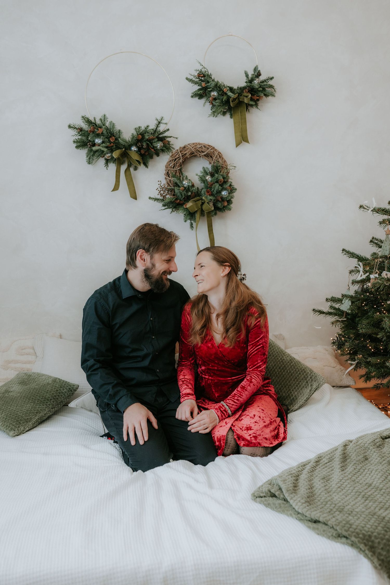 Couple sitting on a white bed smiling at each other with Christmas wreaths on the wall behind them.