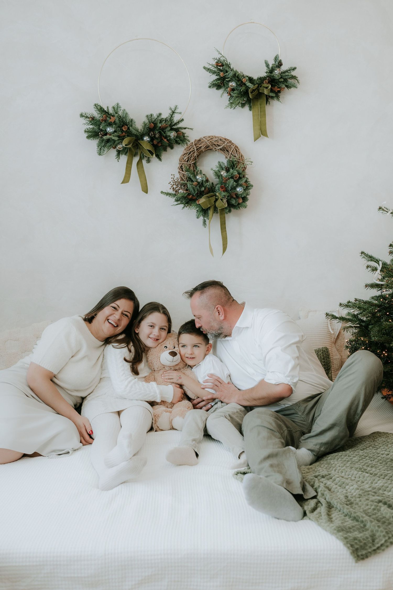 Family of four in white outfits sitting on bed with Christmas wreaths and tree in festive holiday photo session.