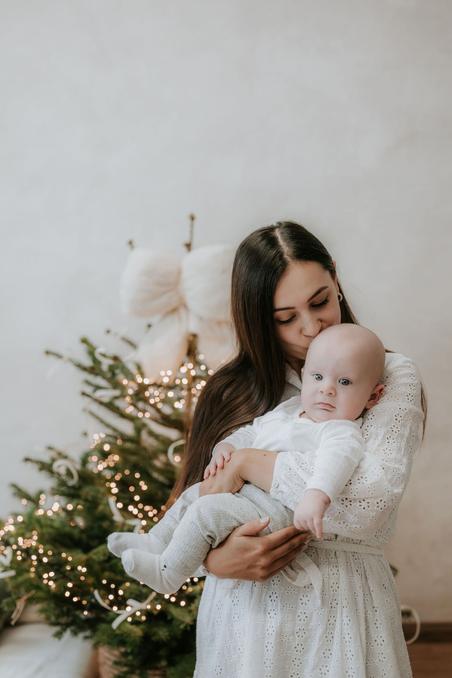 A young mother in a white dress kisses her baby near a decorated Christmas tree with warm glowing lights.