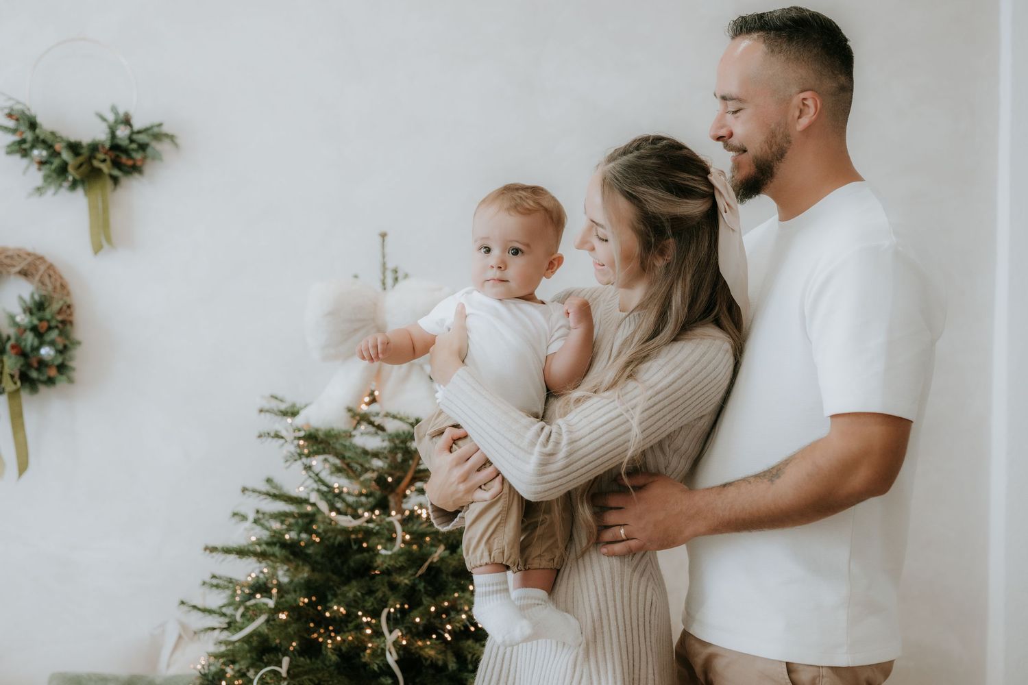 A happy young family with a baby poses near a decorated Christmas tree in a bright, cozy white room.