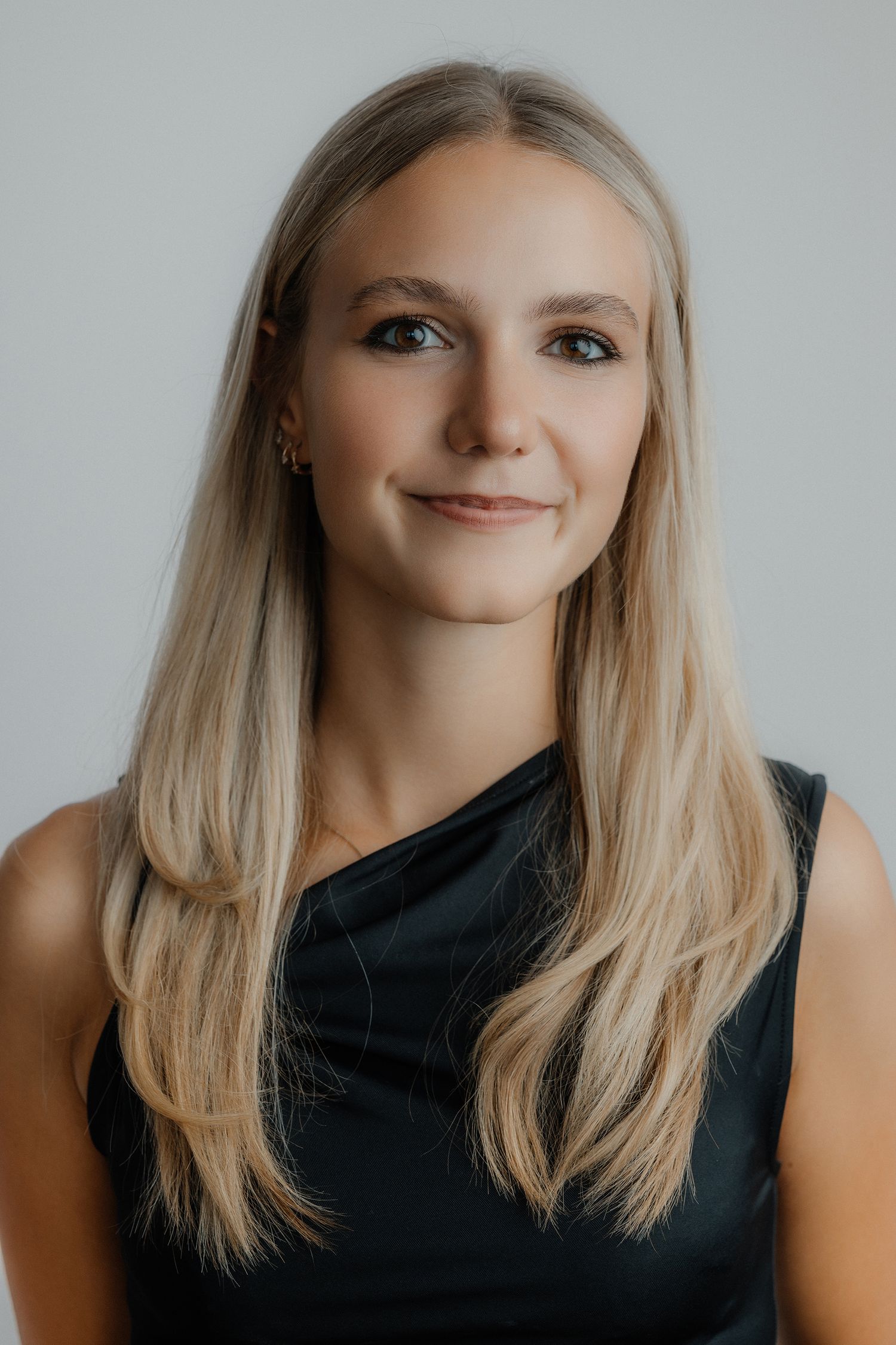 Young blonde woman smiling in a black sleeveless top against a light gray background, professional headshot.