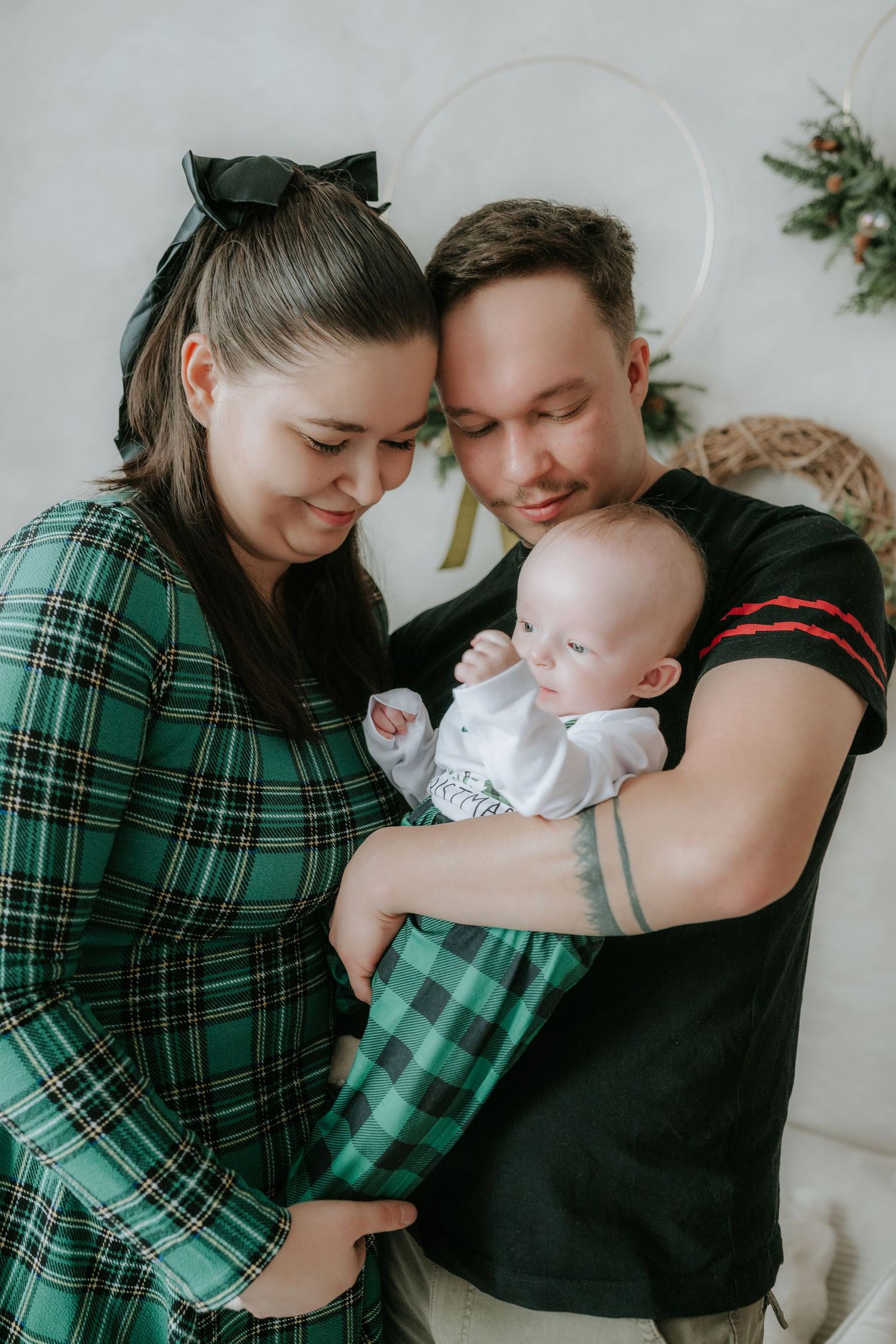 A young family in matching green plaid outfits tenderly holds their newborn baby during a Christmas photo session.
