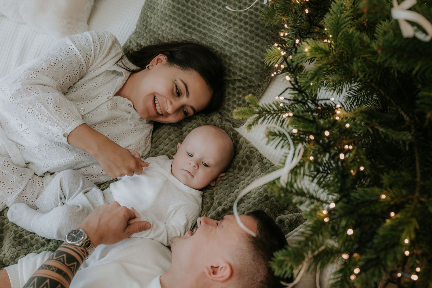 A young family with a baby lies beside a decorated Christmas tree with warm fairy lights on a cozy green blanket.