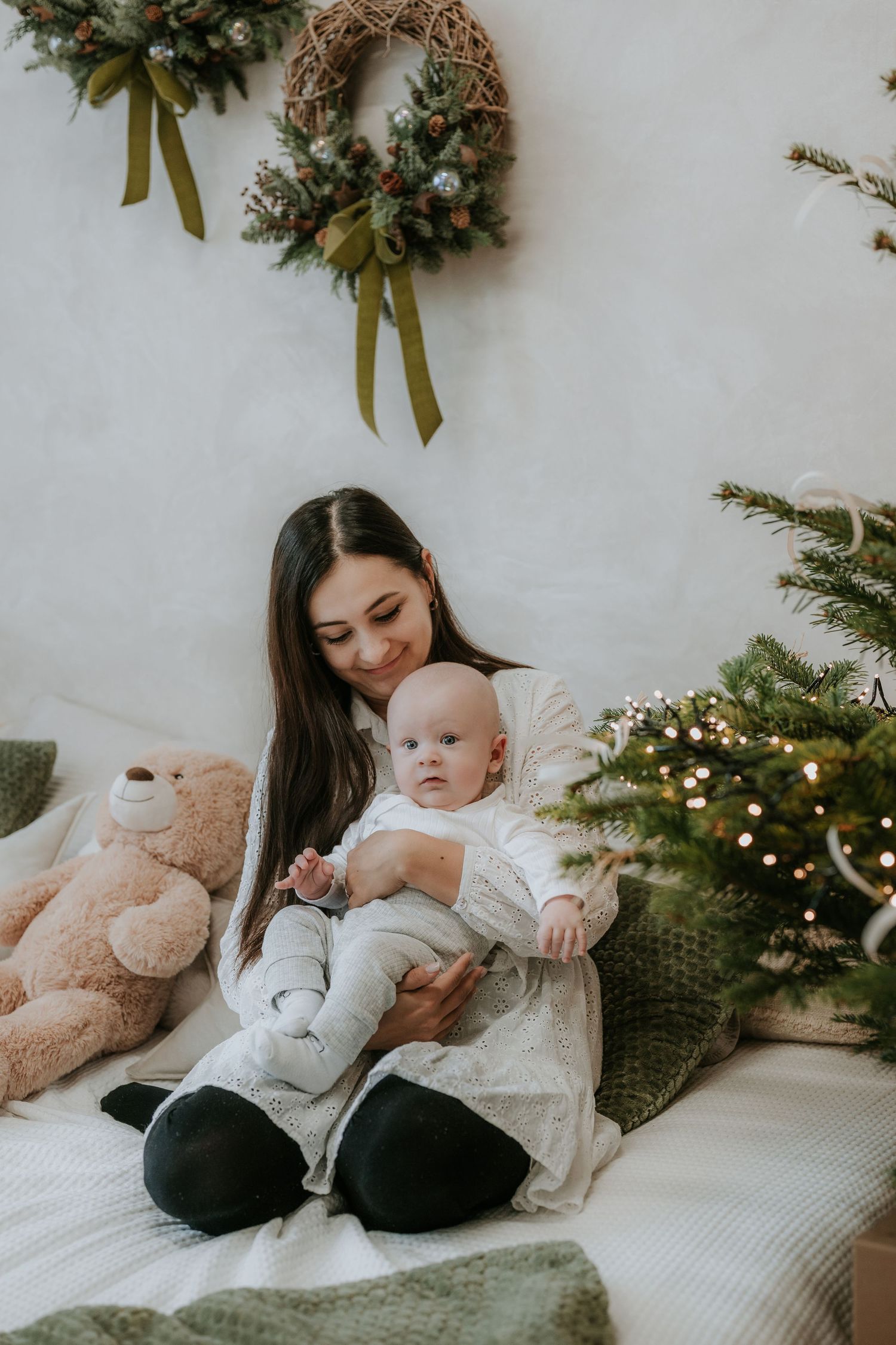 A young mother sits on a bed holding her baby near a decorated Christmas tree and wreath.