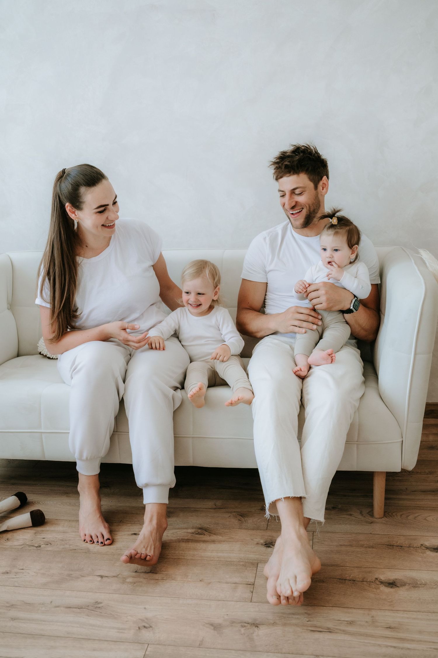 Happy young family with two babies sitting together on a white couch in a bright, cozy living room.