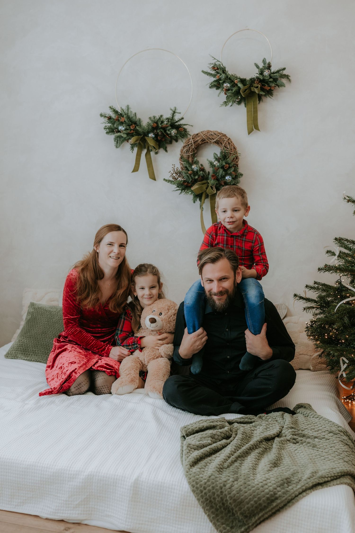 Family of four poses on a white bed with Christmas wreaths and a decorated tree in a festive holiday photo.