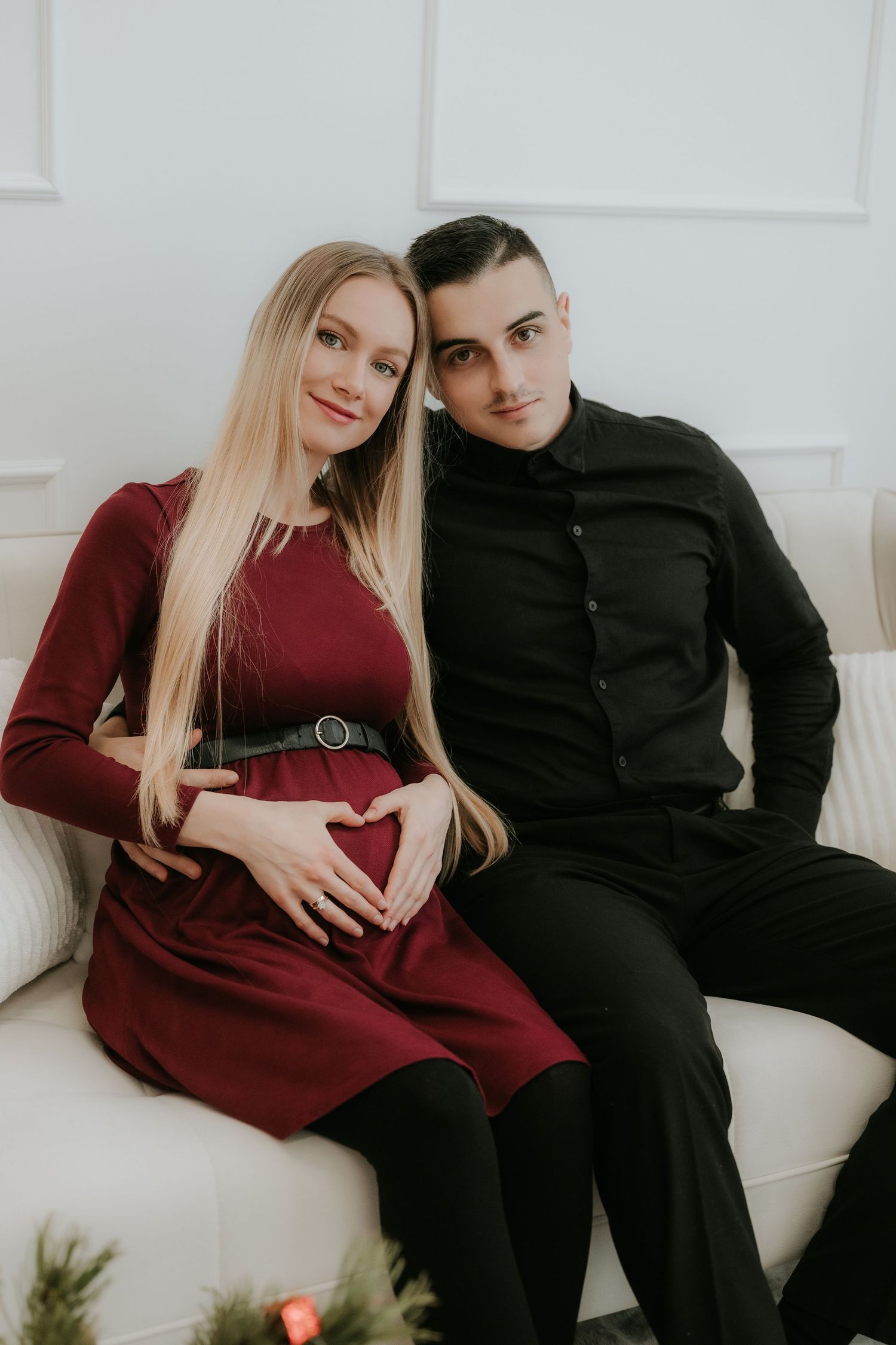 Pregnant couple sitting on white sofa, woman in burgundy dress forming heart shape on baby bump, man in black shirt beside her.