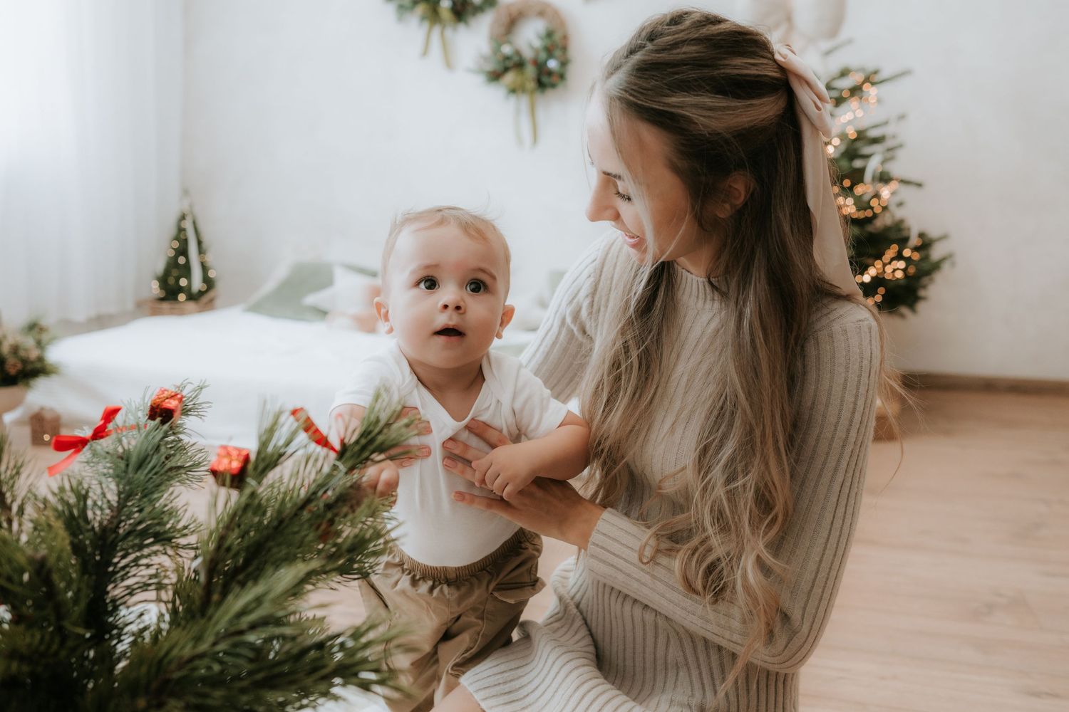 A young mother holds her baby near a Christmas tree in a cozy decorated bedroom during the holiday season.