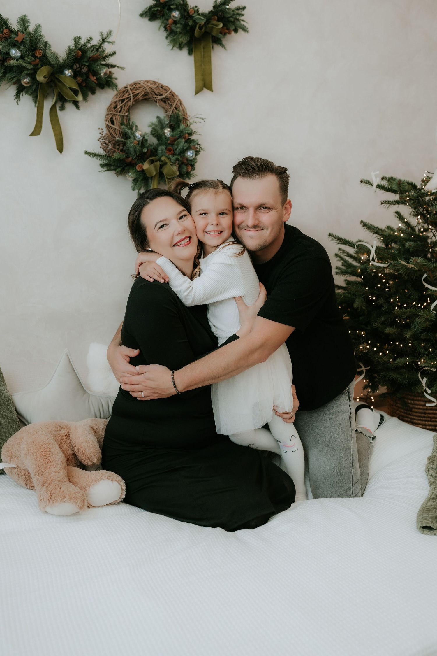 Family of three sharing a warm Christmas hug beside a decorated tree and holiday wreaths.