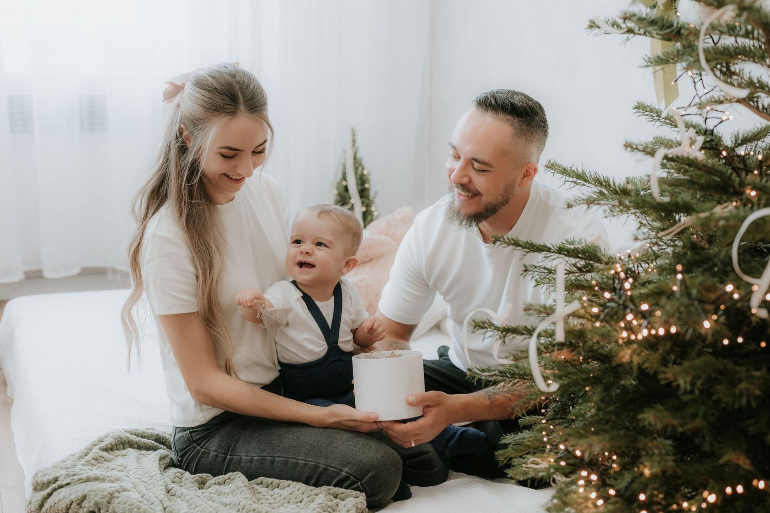 Happy family with baby celebrating Christmas near decorated tree, exchanging gifts in bright white room.