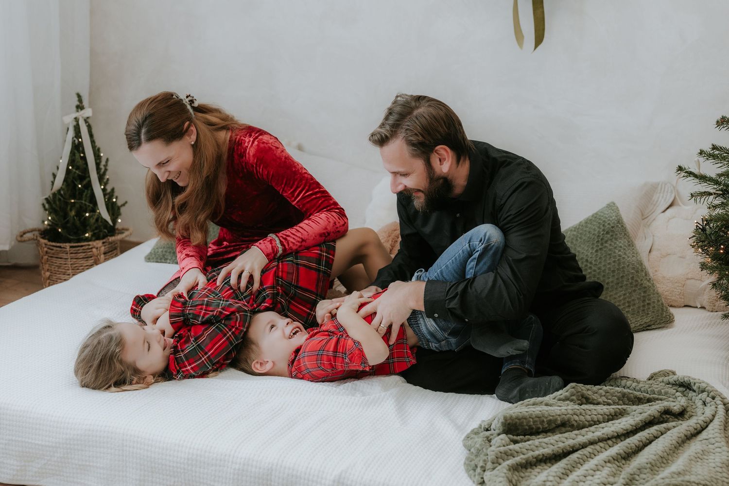 Family in Christmas pajamas playing on a white bed with holiday trees in the background.