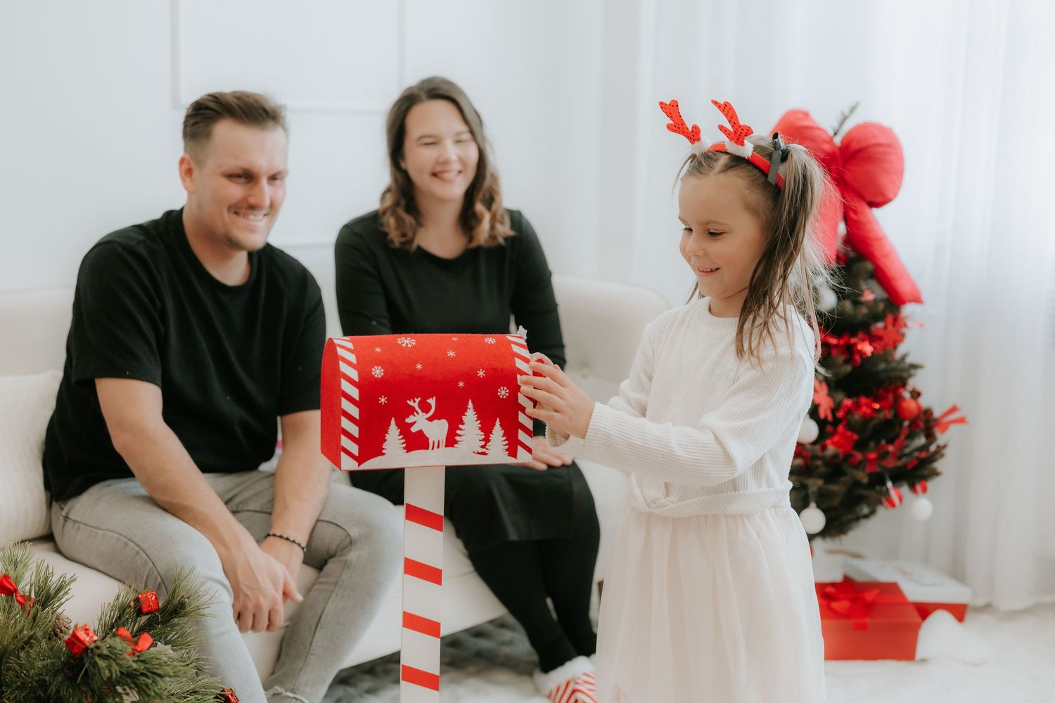 A young girl in reindeer antlers opens a Christmas gift while her smiling parents watch from a white sofa near a decorated tree.