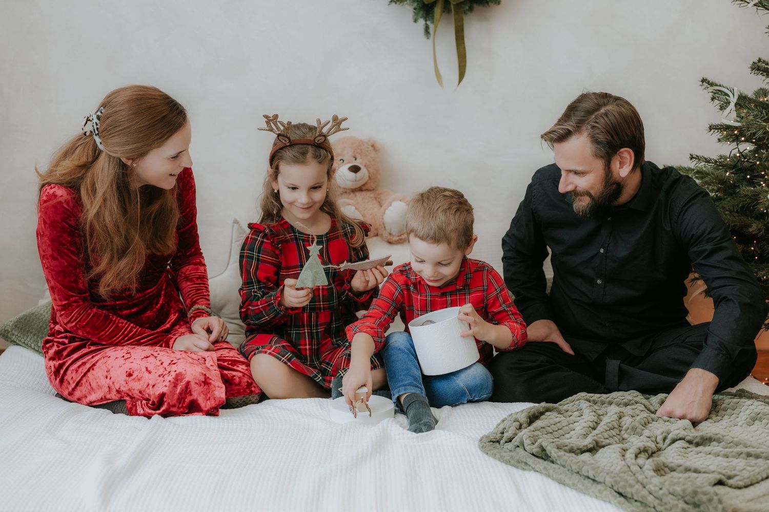 Family of four in holiday pajamas sitting together on a white bed near a Christmas tree, sharing a joyful moment.