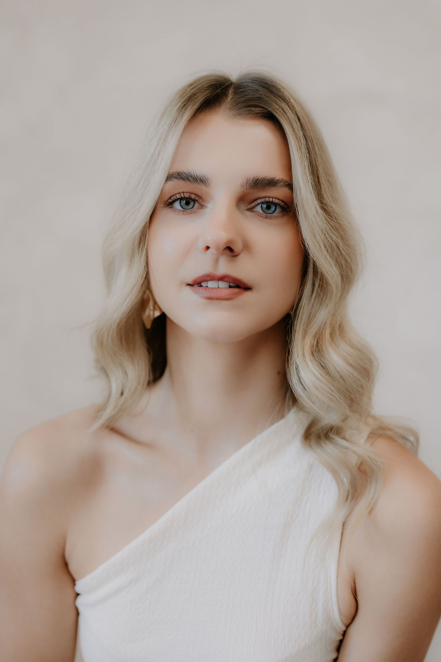 Young woman with blonde wavy hair wearing a white one-shoulder top against a neutral beige background.