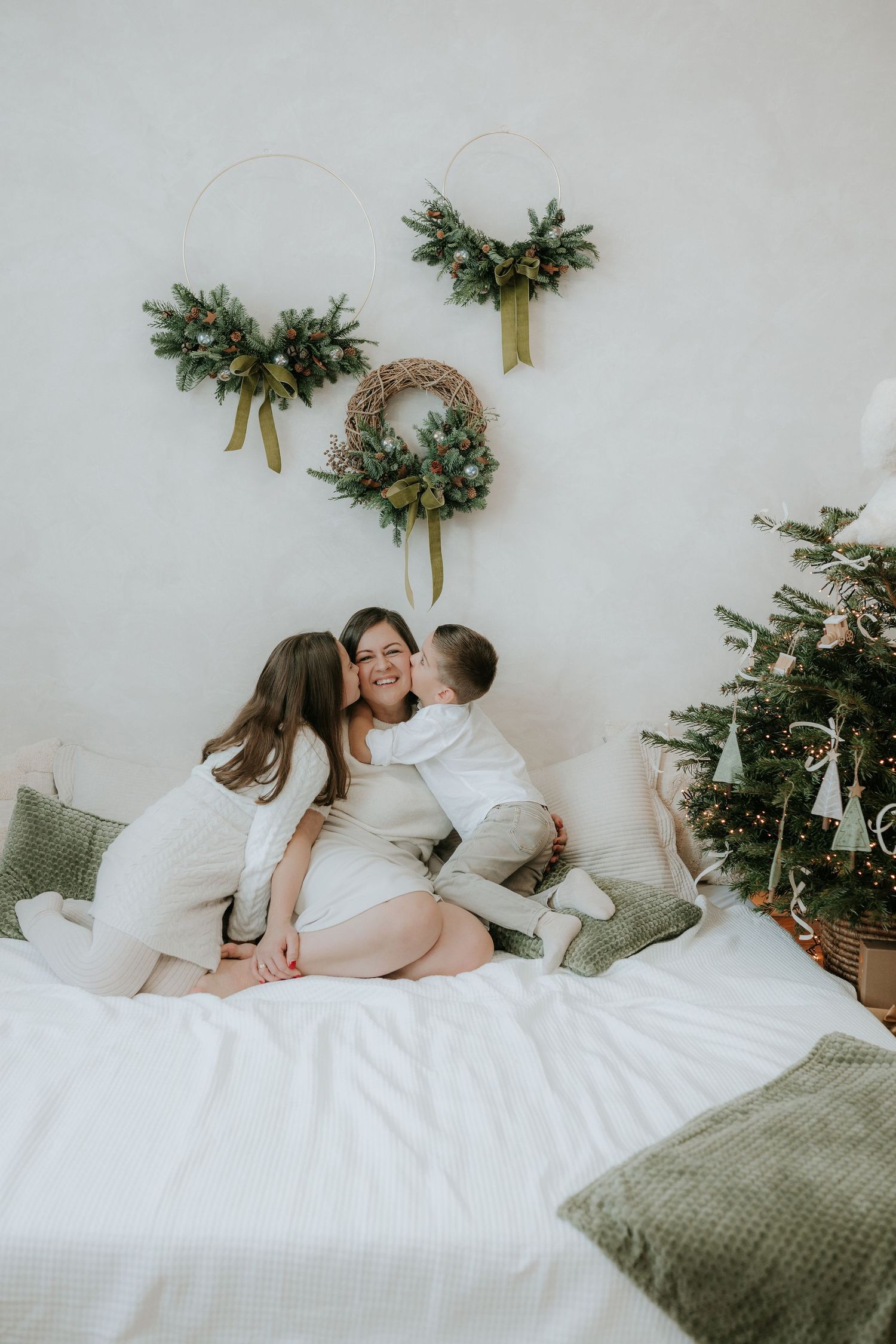 Mother sitting on bed receiving kisses from two children near Christmas tree with holiday wreaths on wall.