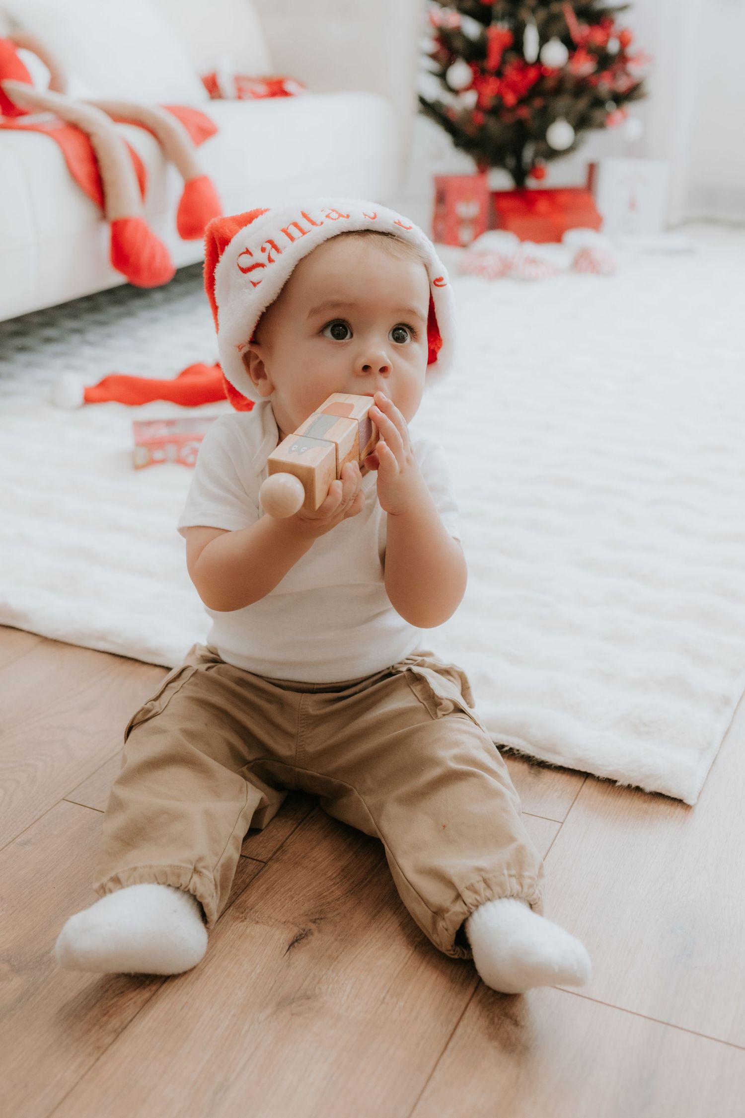 Baby in Santa hat sits on floor chewing wooden toy near Christmas tree with gifts.
