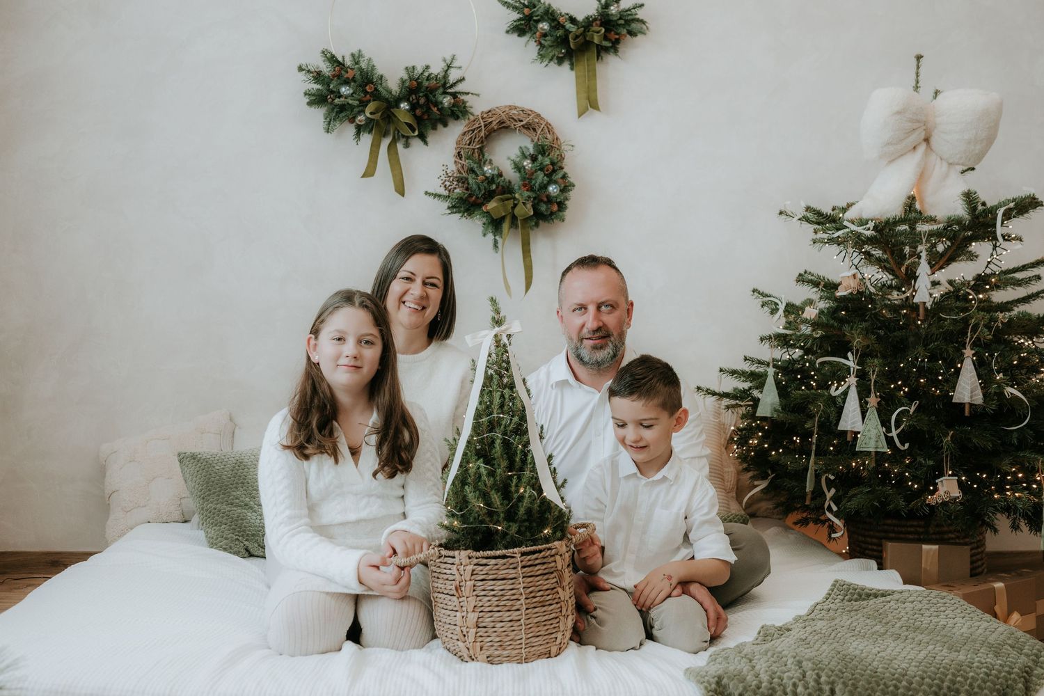 Family of four in white outfits posing with a small Christmas tree and holiday wreaths in a festive decorated room.