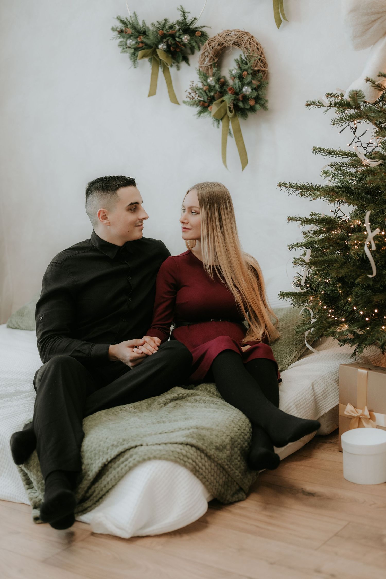 Couple sitting together on a bed near a decorated Christmas tree, holding hands and smiling at each other.