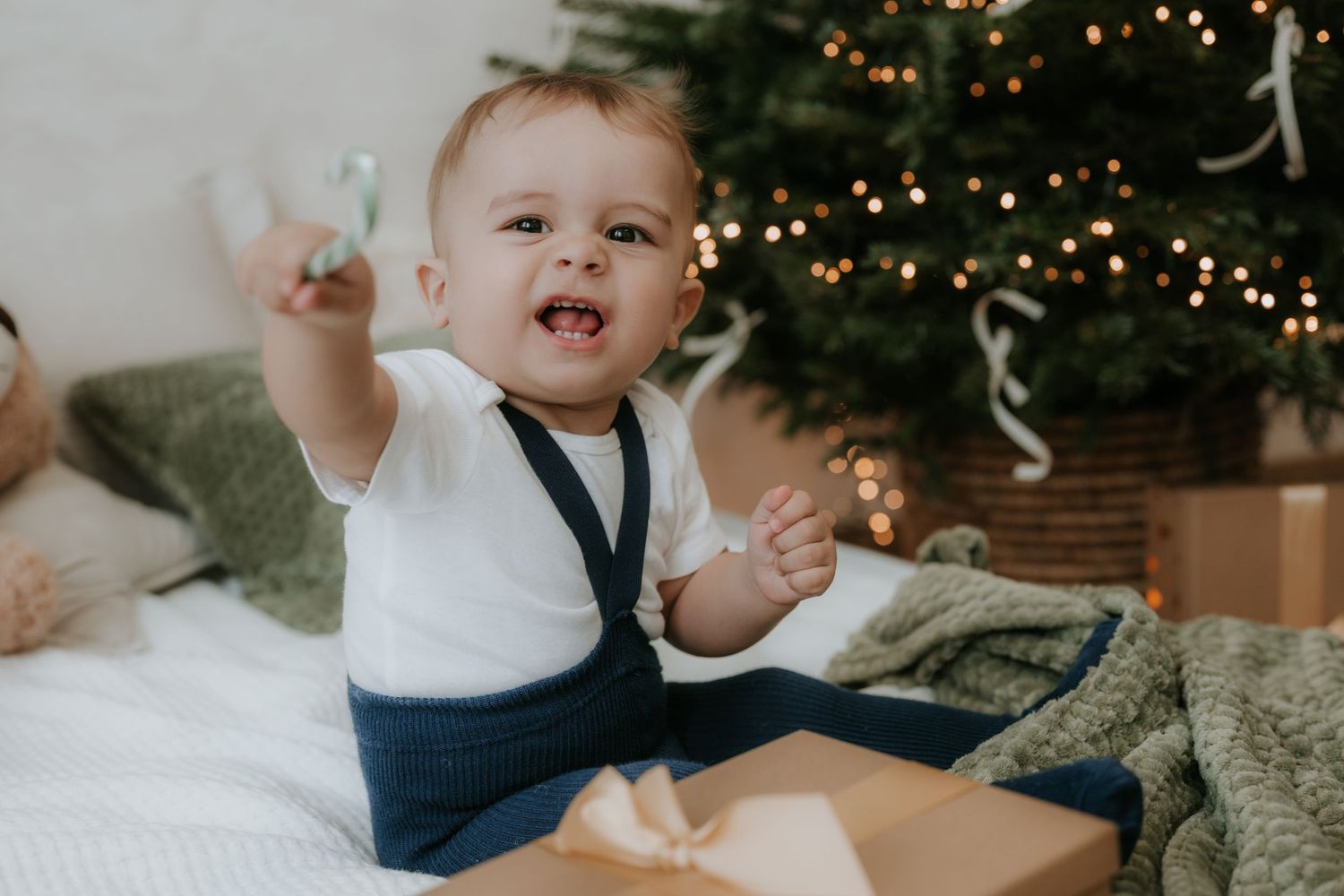 Happy baby in white shirt sits by Christmas tree with gift box, reaching out and smiling joyfully.