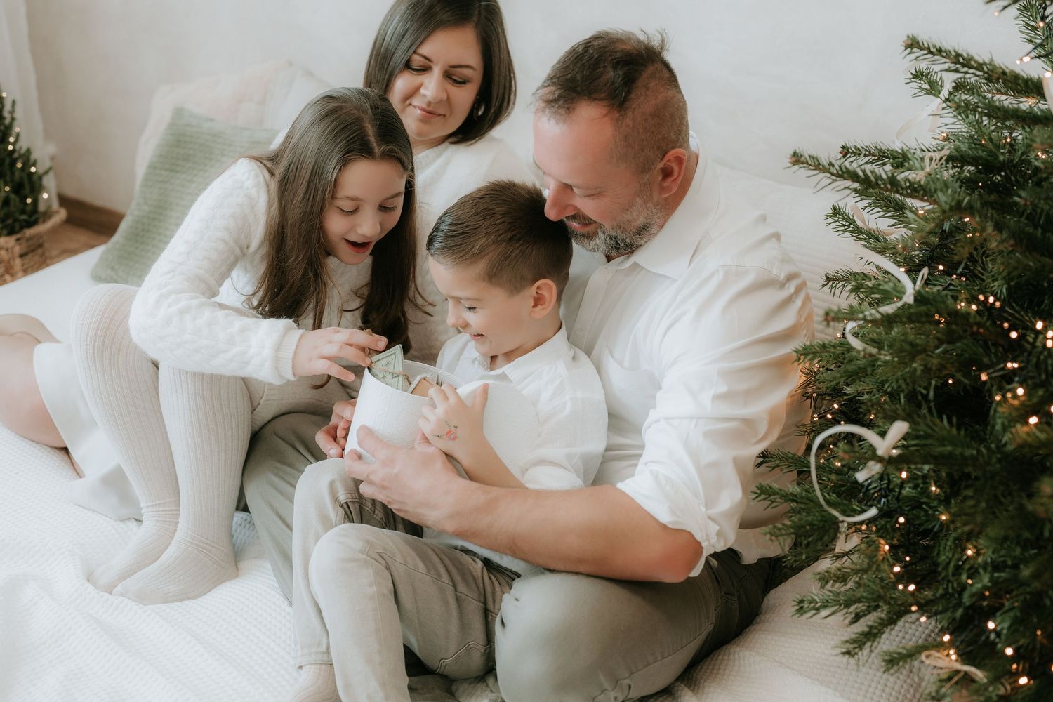 Family of four in white outfits opening a Christmas gift together beside a decorated tree.