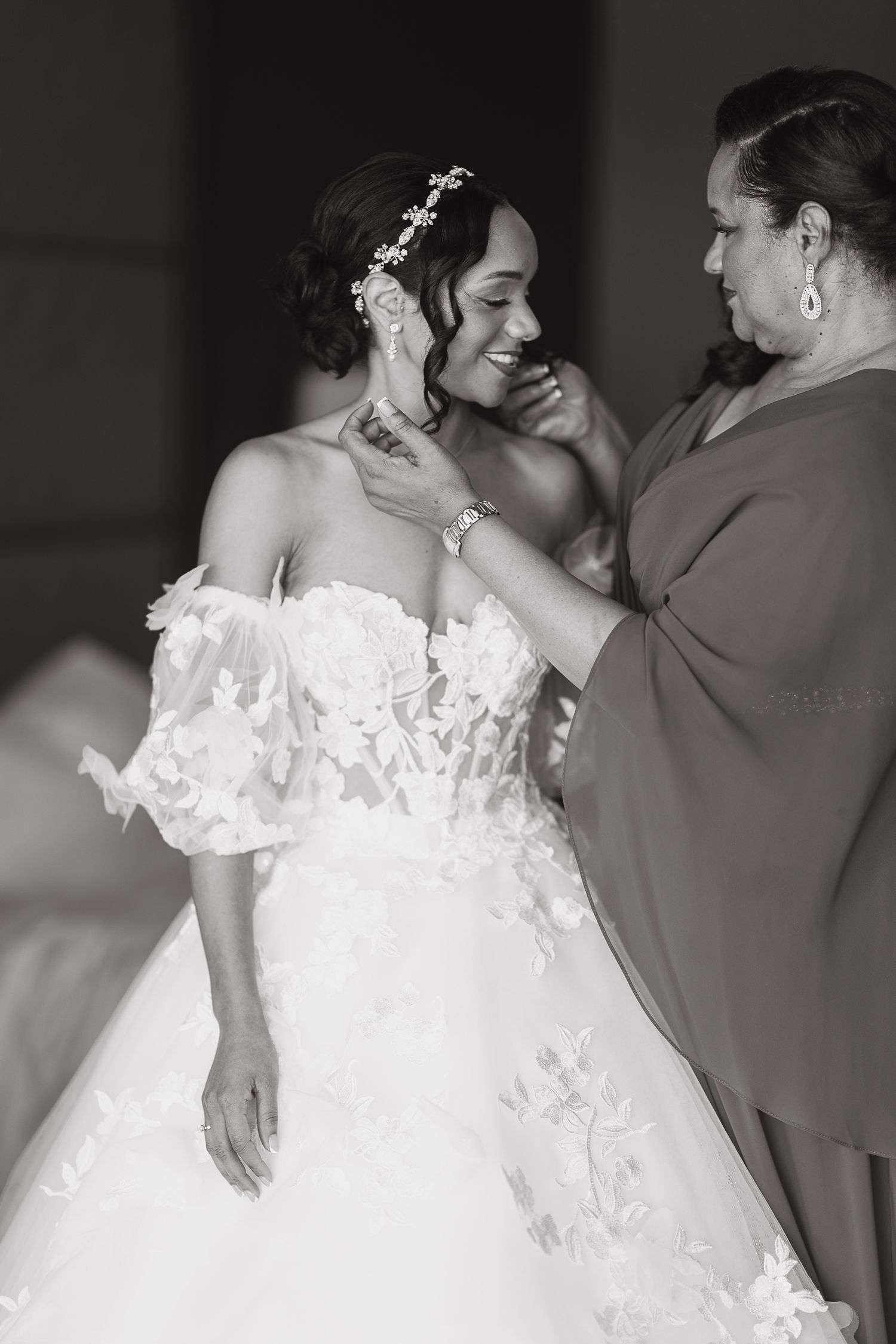 Black and white photo of mother adjusting brides hair accessory as she smiles in floral wedding gown.