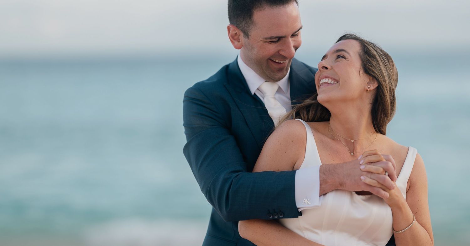Happy couple embracing on a beach, groom in navy suit and bride in white dress, with ocean waves in the background.