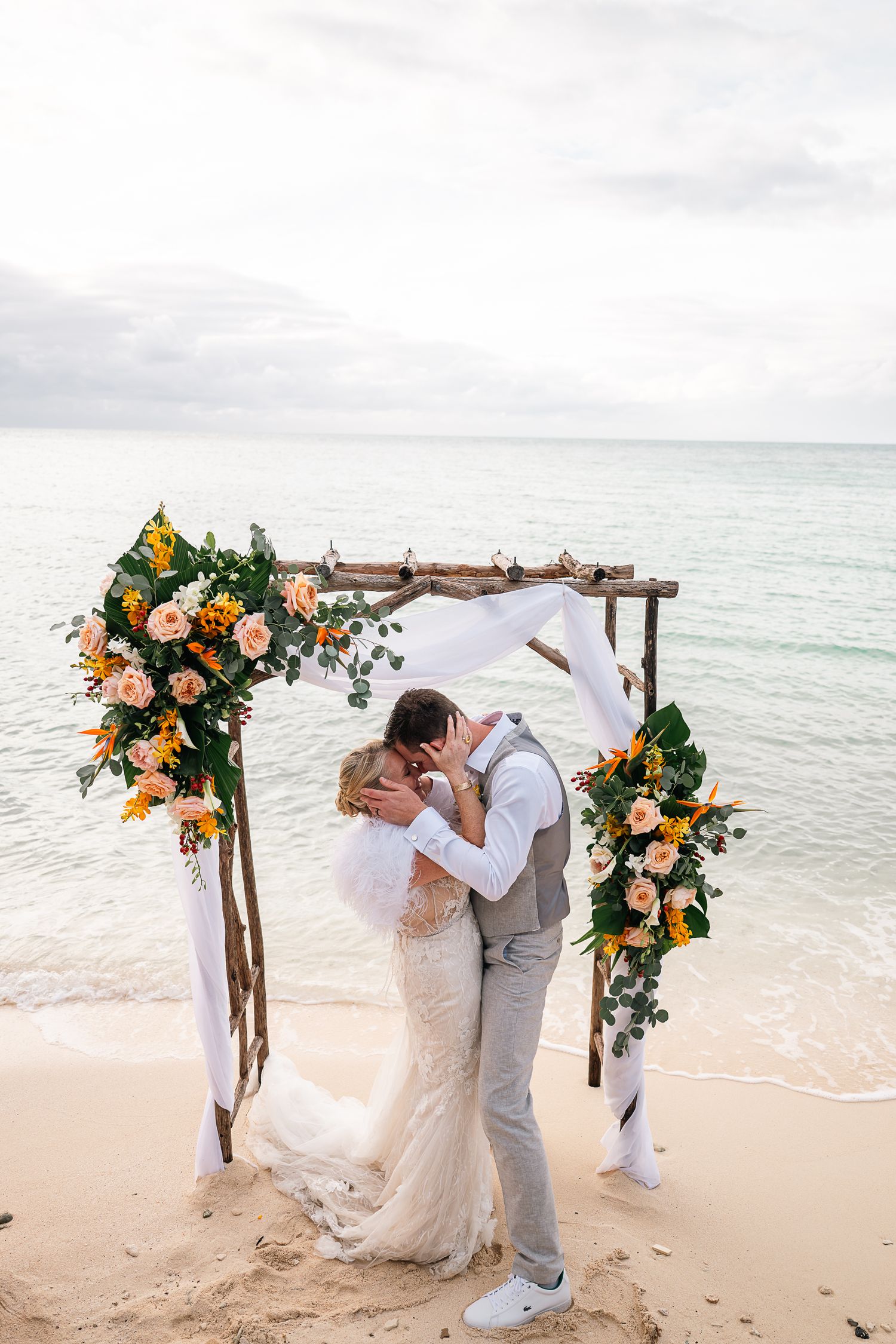 Bride and groom share first kiss under floral arch decorated with sunflowers and roses on a tropical beach.