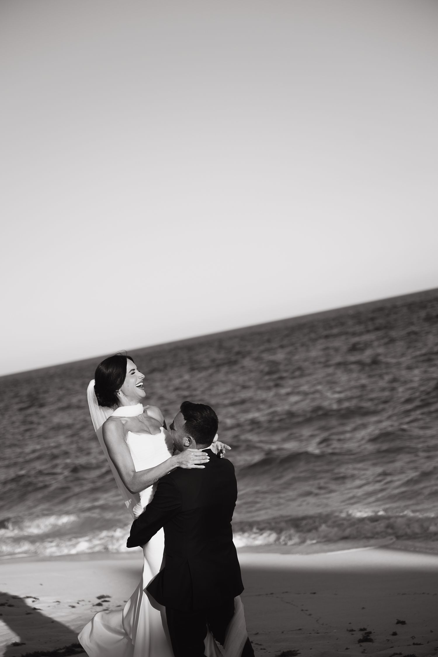 Black and white photo of bride and groom embracing on beach, smiling at each other with ocean waves behind them.