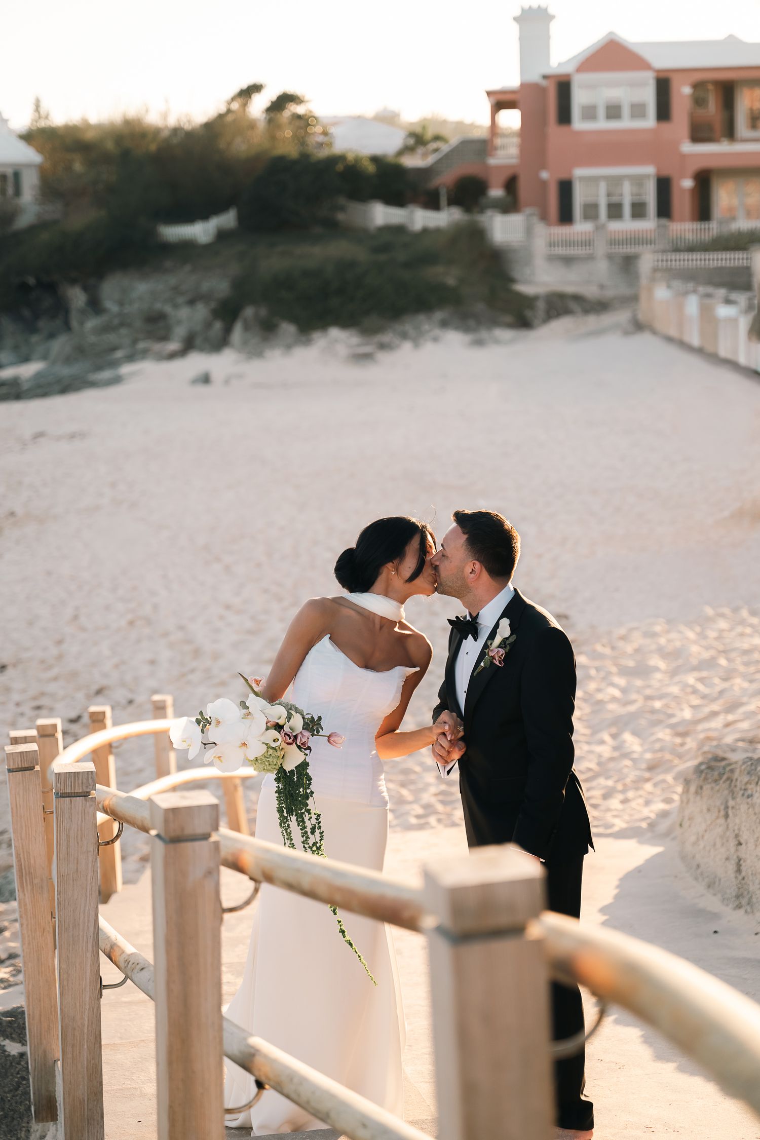 Bride and groom kissing on wooden beach boardwalk at golden hour, bride holding bouquet with sandy dunes behind.