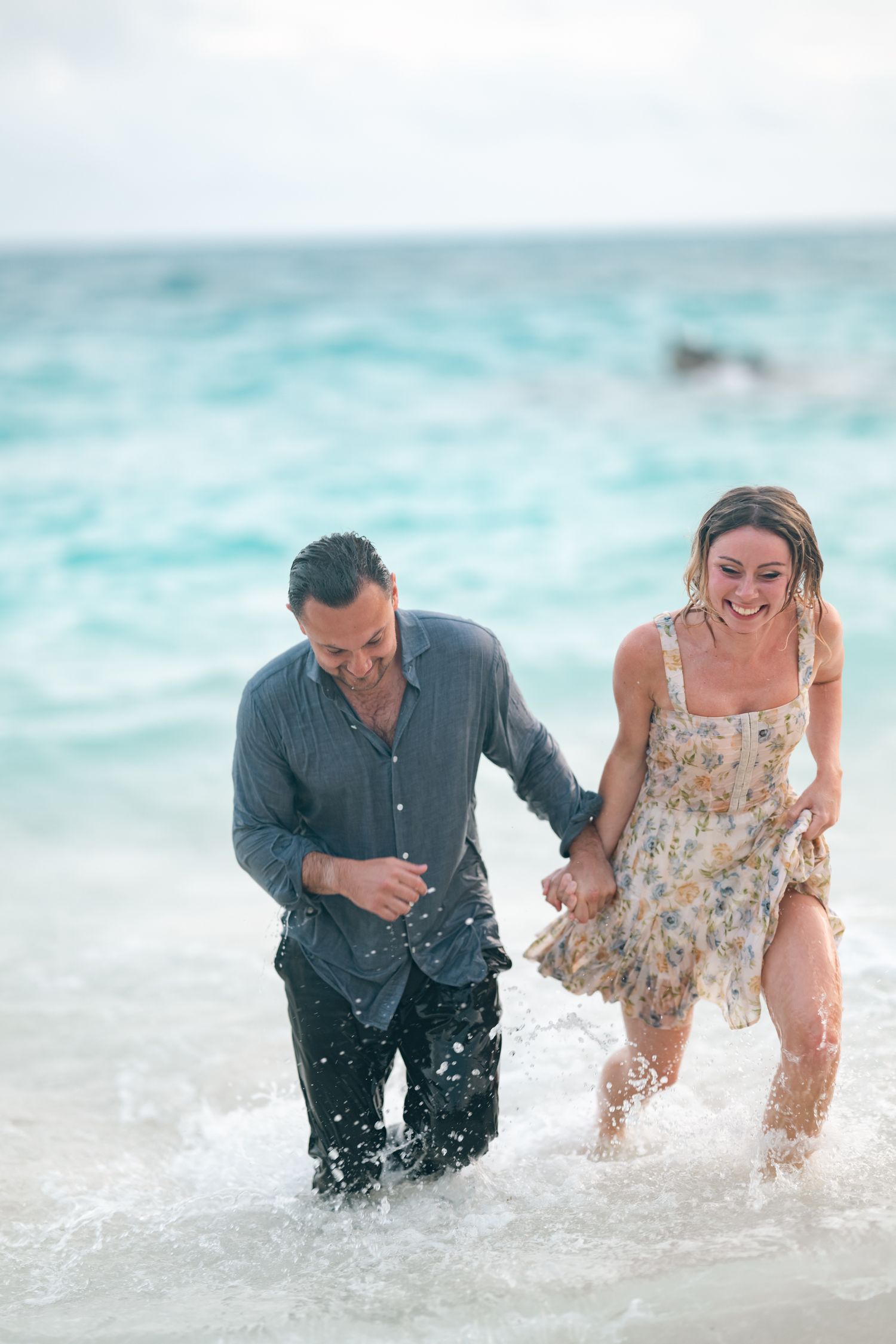 Couple laughing and holding hands while walking through ocean waves on a tropical beach.