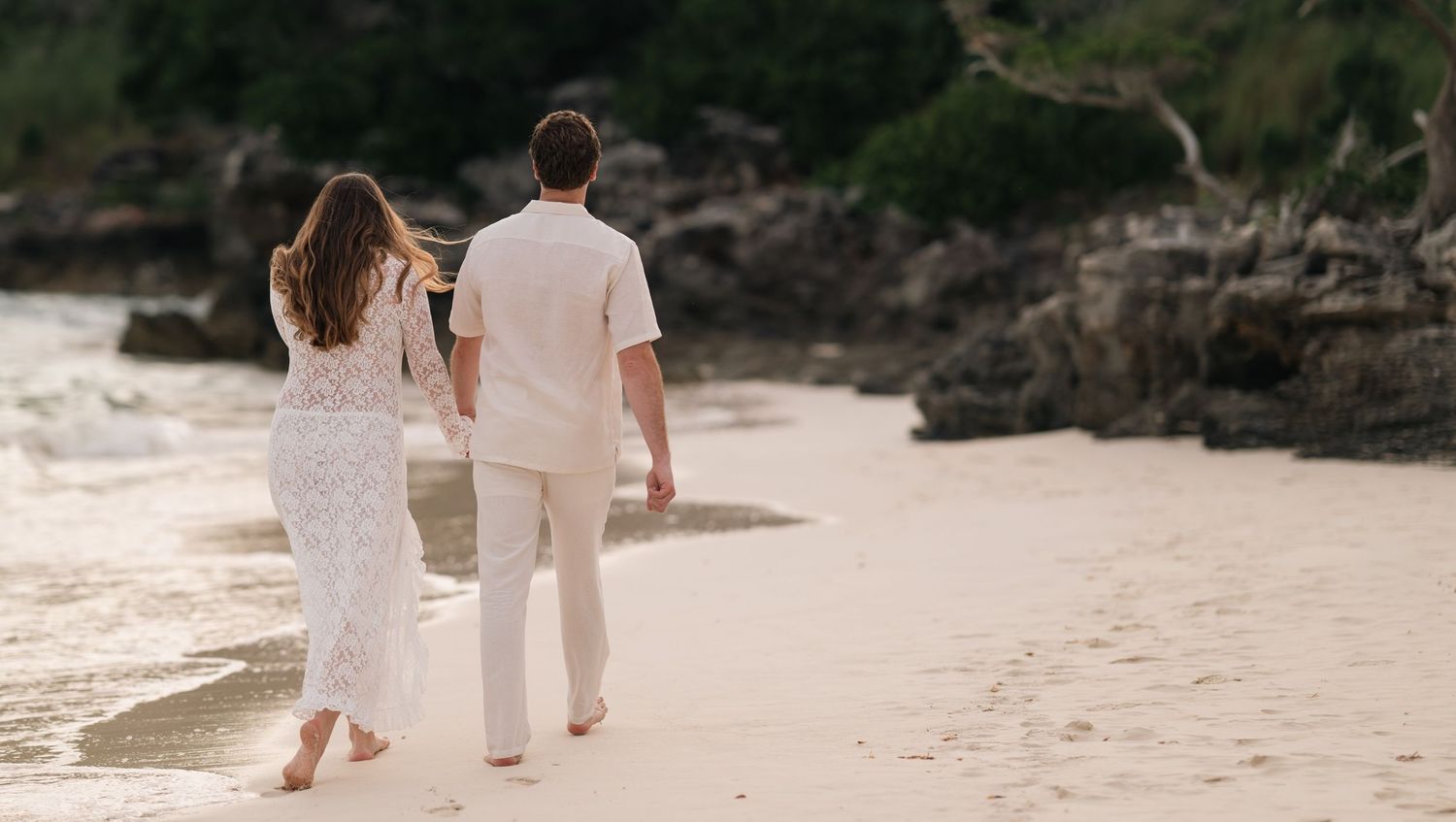 Couple in white outfits walking barefoot on a sandy tropical beach near rocky shores at sunset.