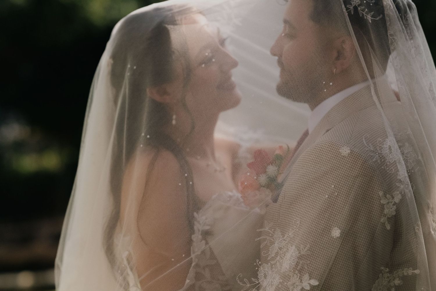Bride and groom sharing an intimate moment beneath a flowing lace wedding veil in soft sunlight.