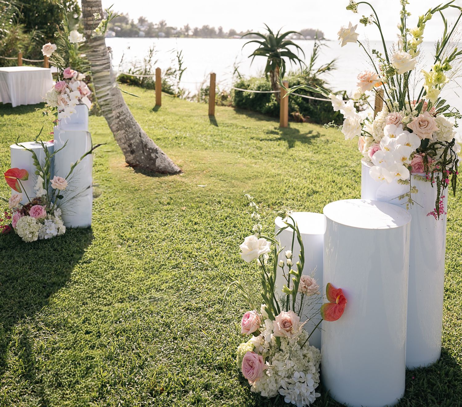 White cylindrical pedestals adorned with pink and white floral arrangements line a lush green lawn wedding aisle by the water.