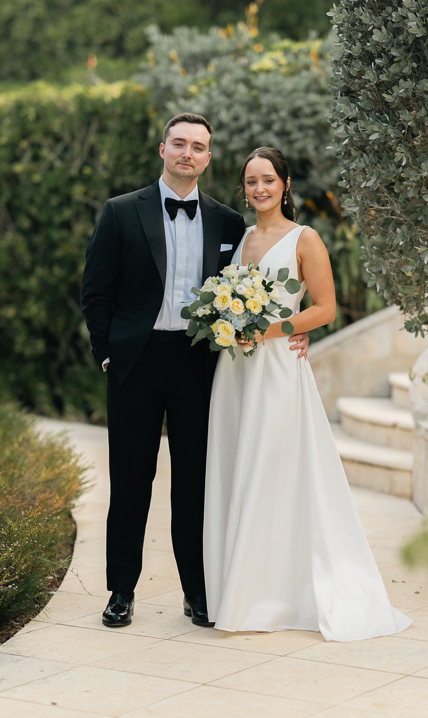 Bride and groom pose together in garden, bride holding yellow and white bouquet.