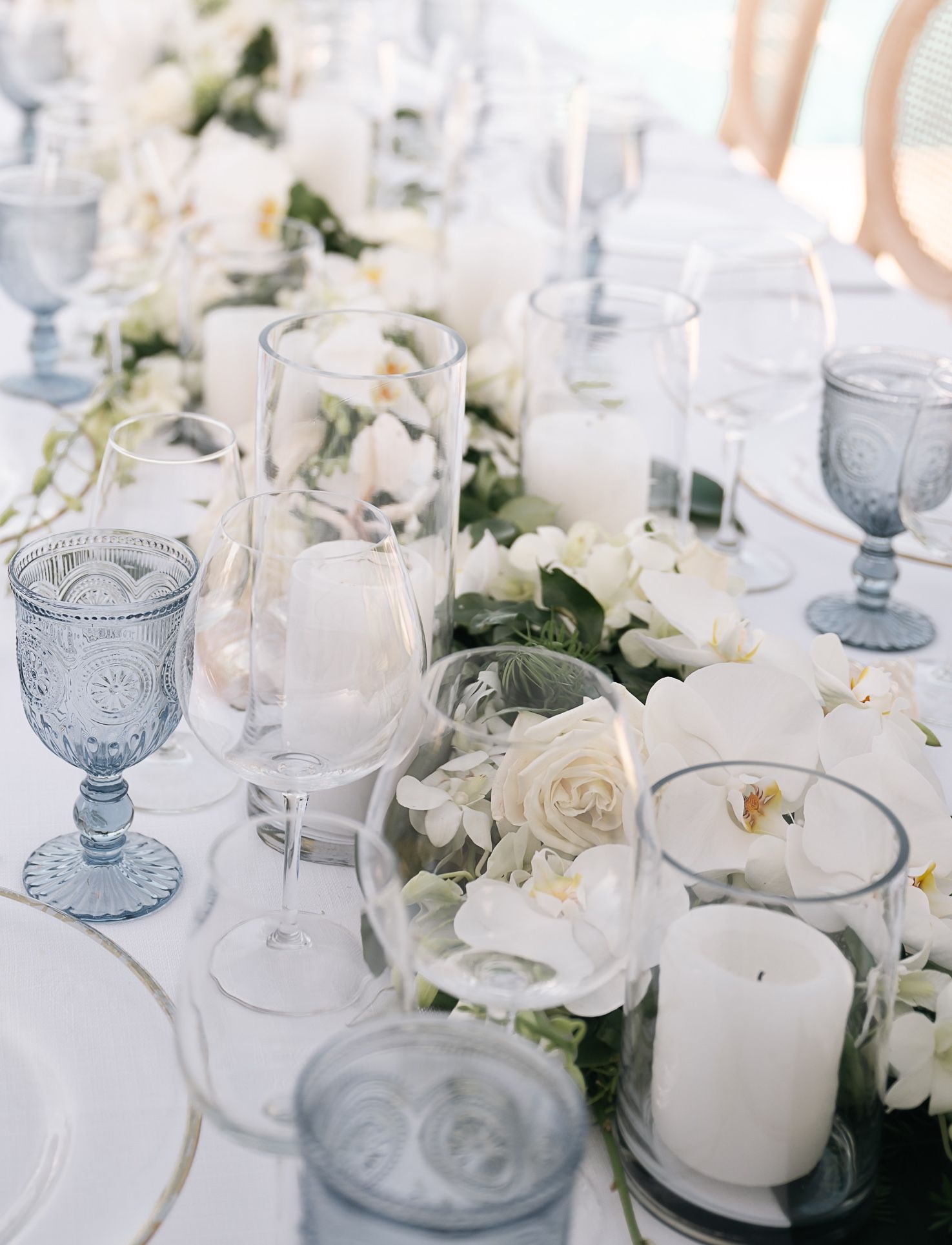 Close-up of wedding tablescape featuring white roses, orchids, blue goblets, and pillar candles.