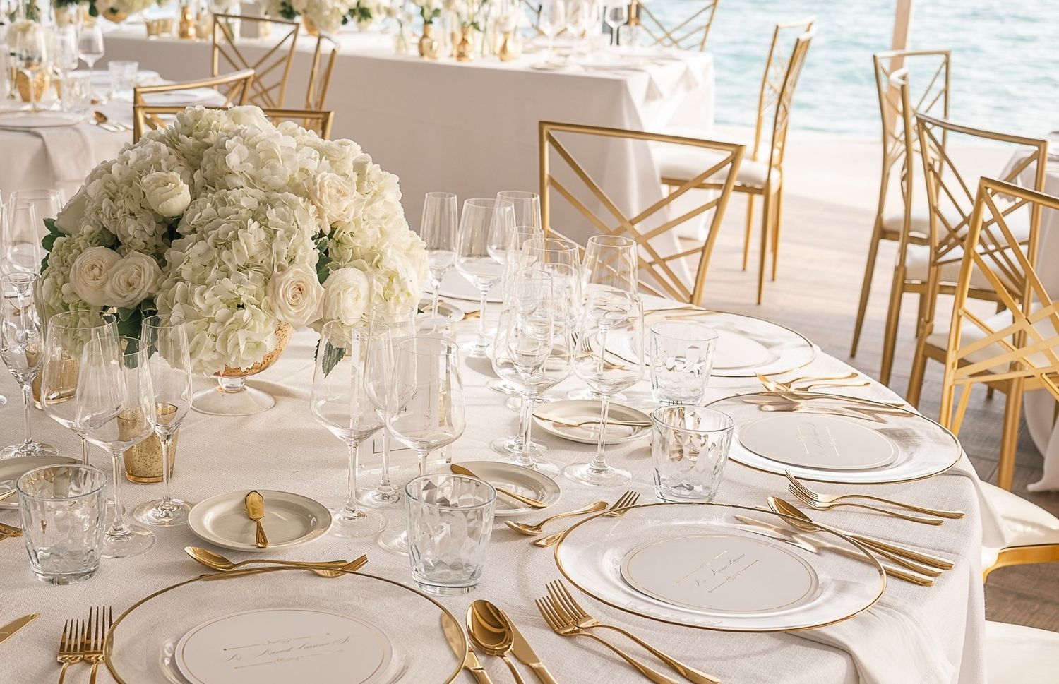 Elegant outdoor wedding reception table with white hydrangea centerpiece, gold accents, and crystal glasses by the ocean.