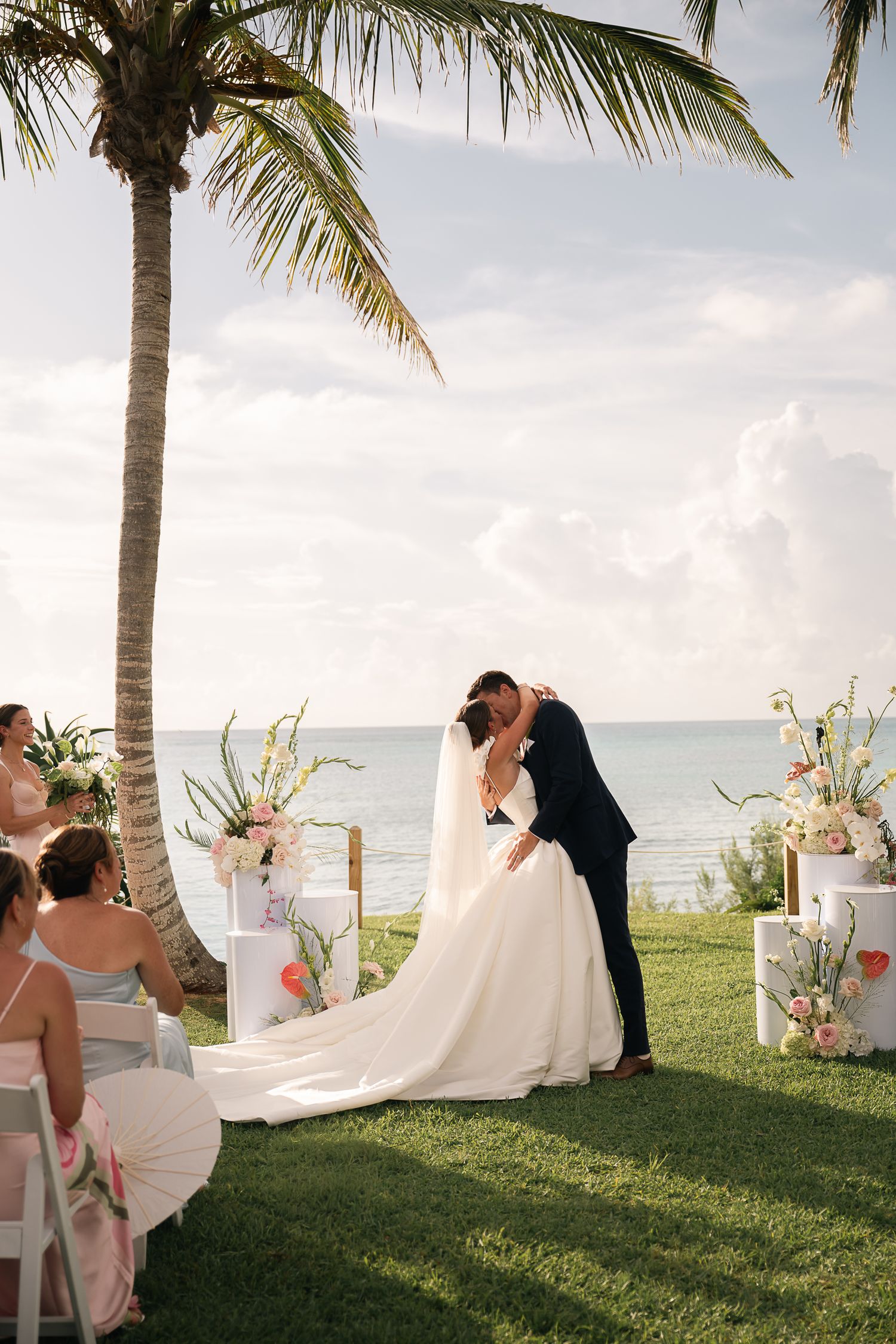Bride and groom share first kiss at romantic beachside wedding ceremony under a tropical palm tree.