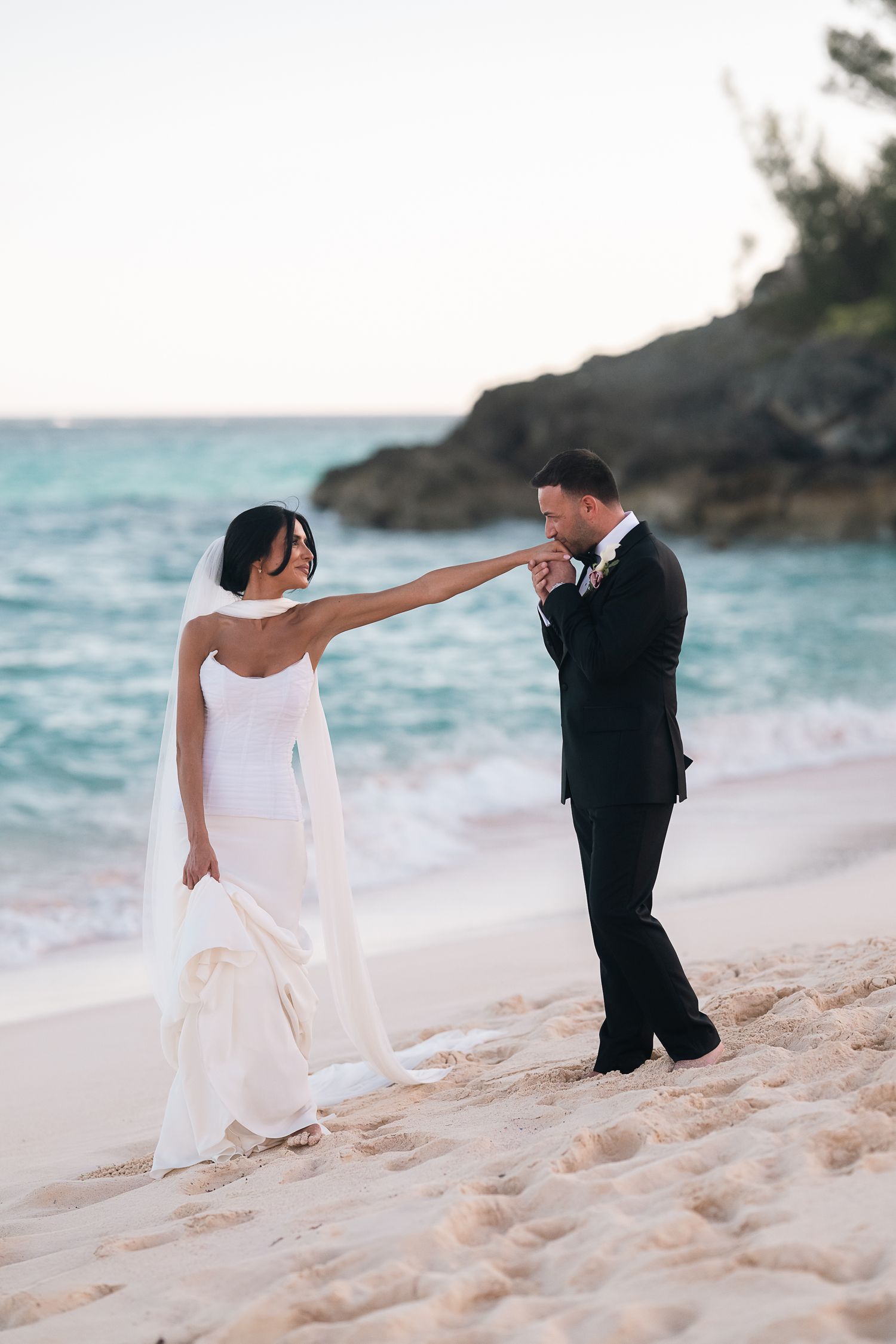 Groom kissing bride's hand on sandy beach at sunset, with turquoise ocean waves and rocky coastline in background.