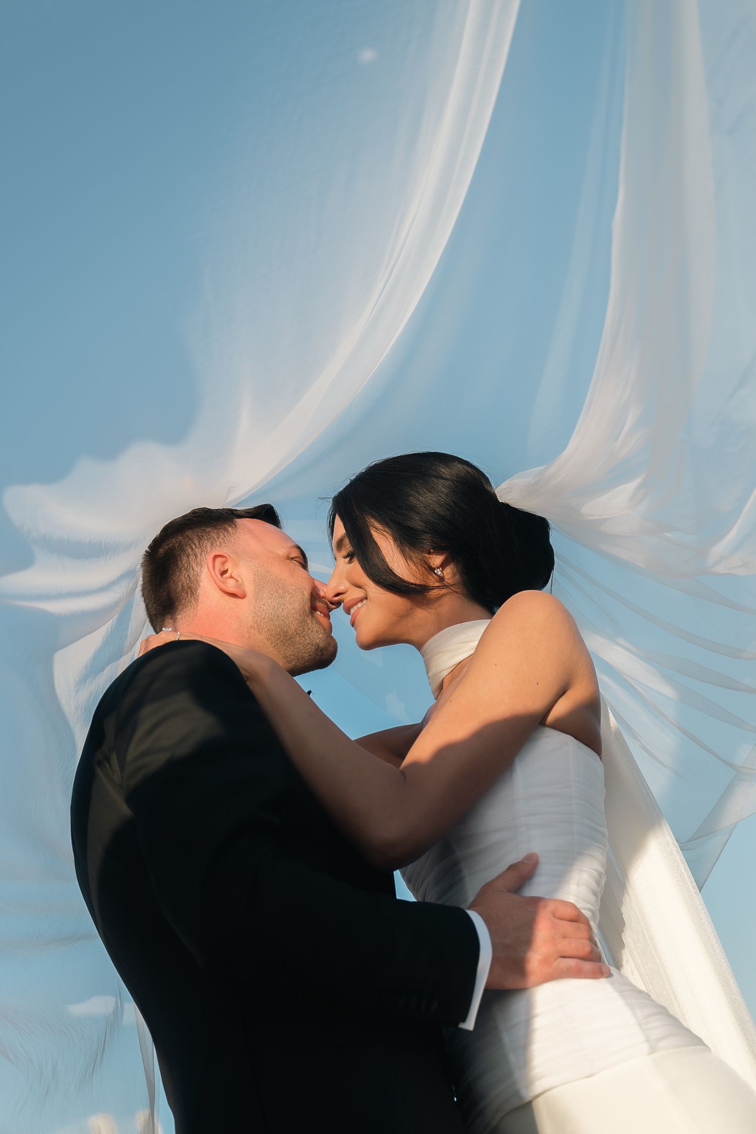 Bride and groom sharing a kiss under flowing white veil against a clear blue sky backdrop.