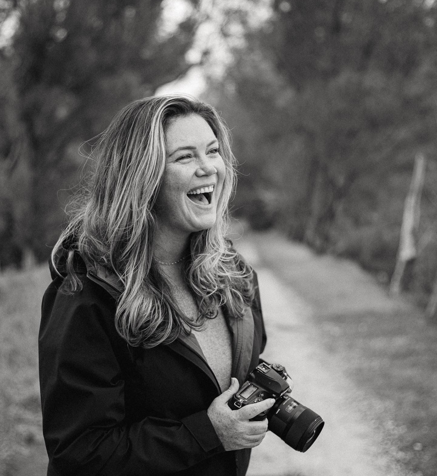 A laughing woman photographer holds a DSLR camera outdoors on a tree-lined path in black and white.