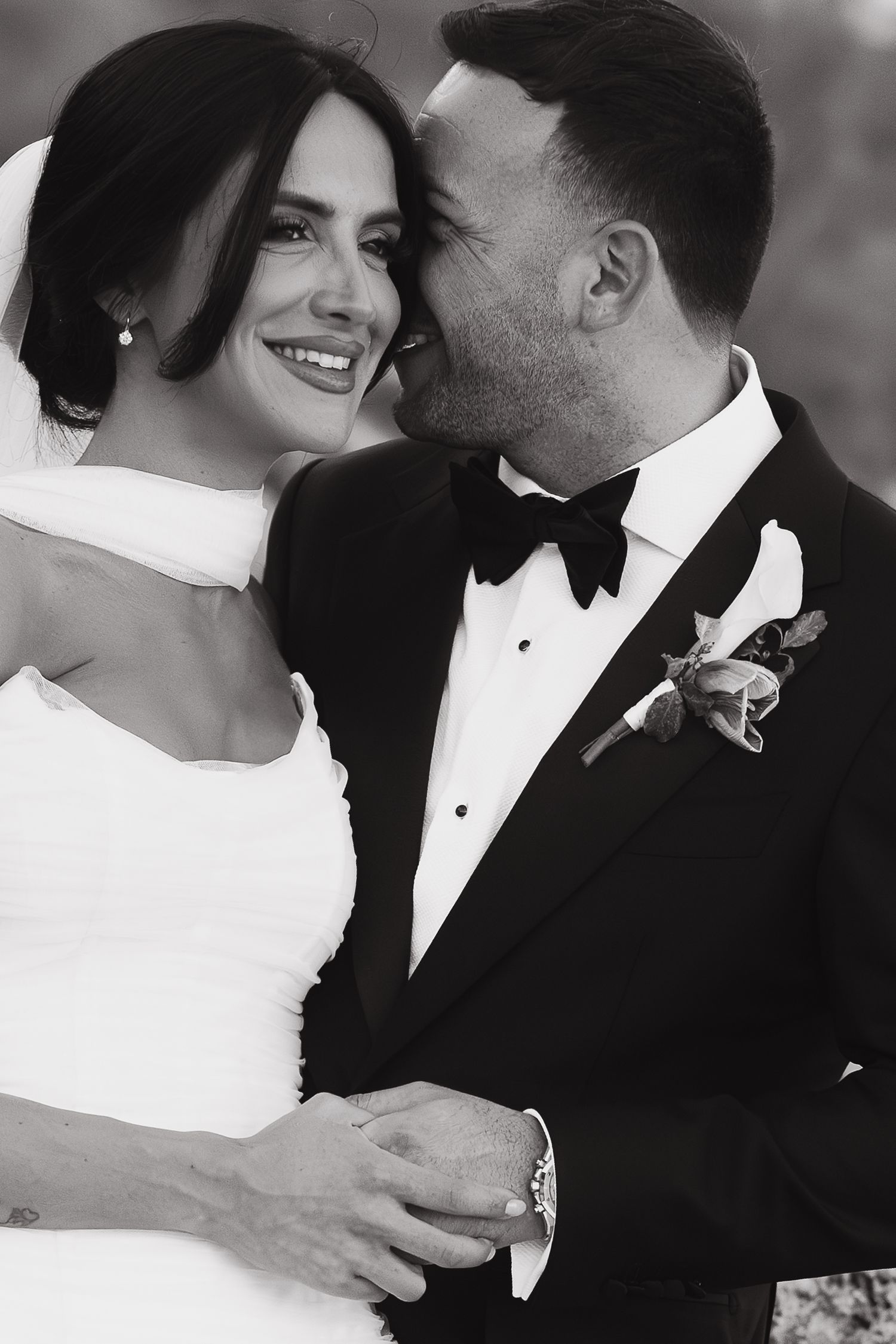 Black and white photo of a smiling bride in white gown and groom in tuxedo sharing a romantic moment.