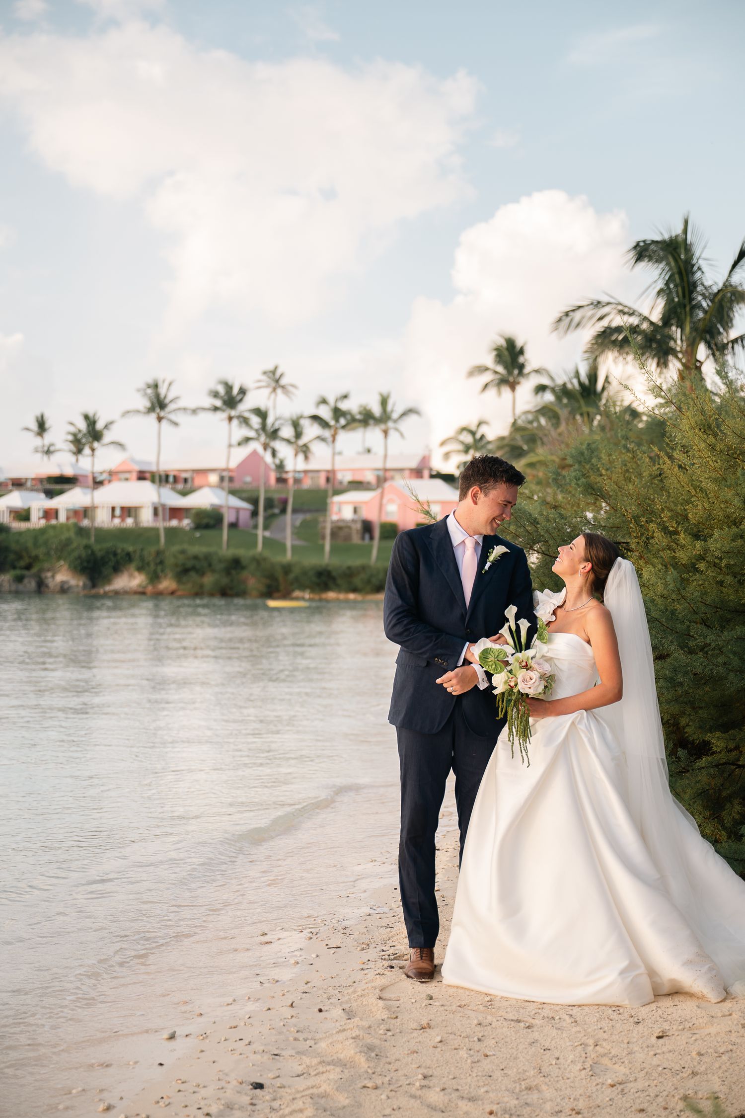 Bride and groom walking along a sandy beach shore with palm trees and pink building backdrop.