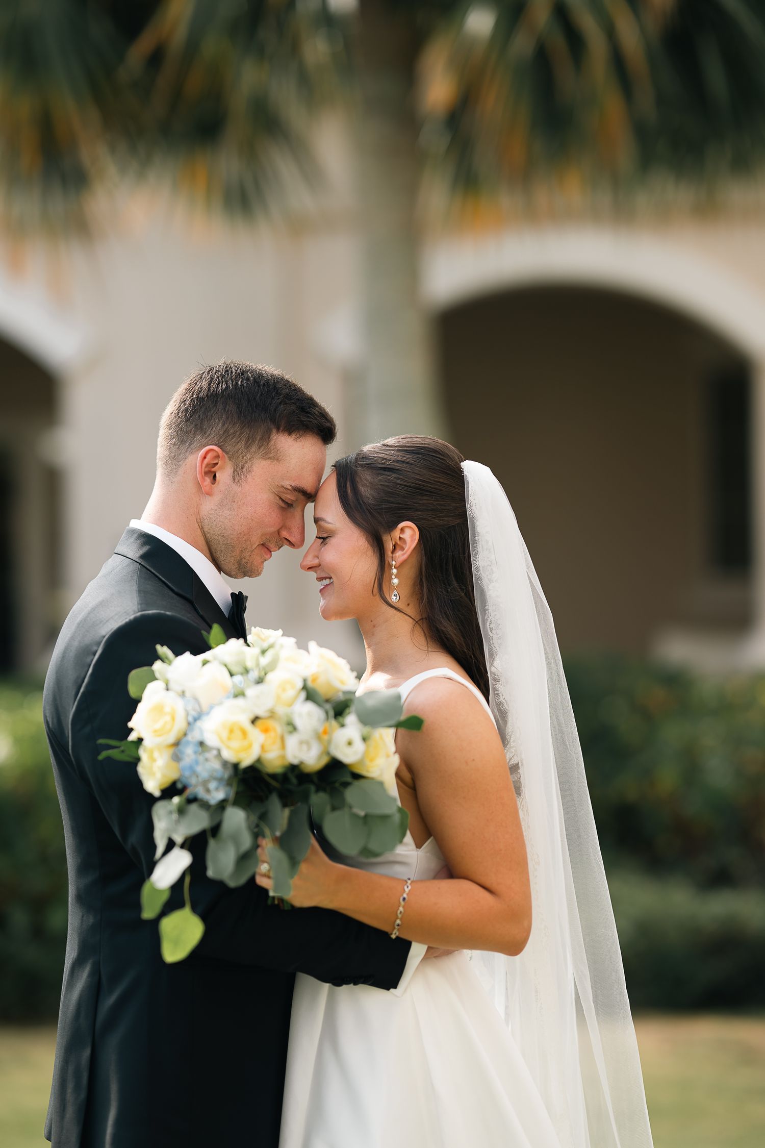 Bride and groom sharing an intimate forehead touch moment outdoors with tropical palm backdrop.