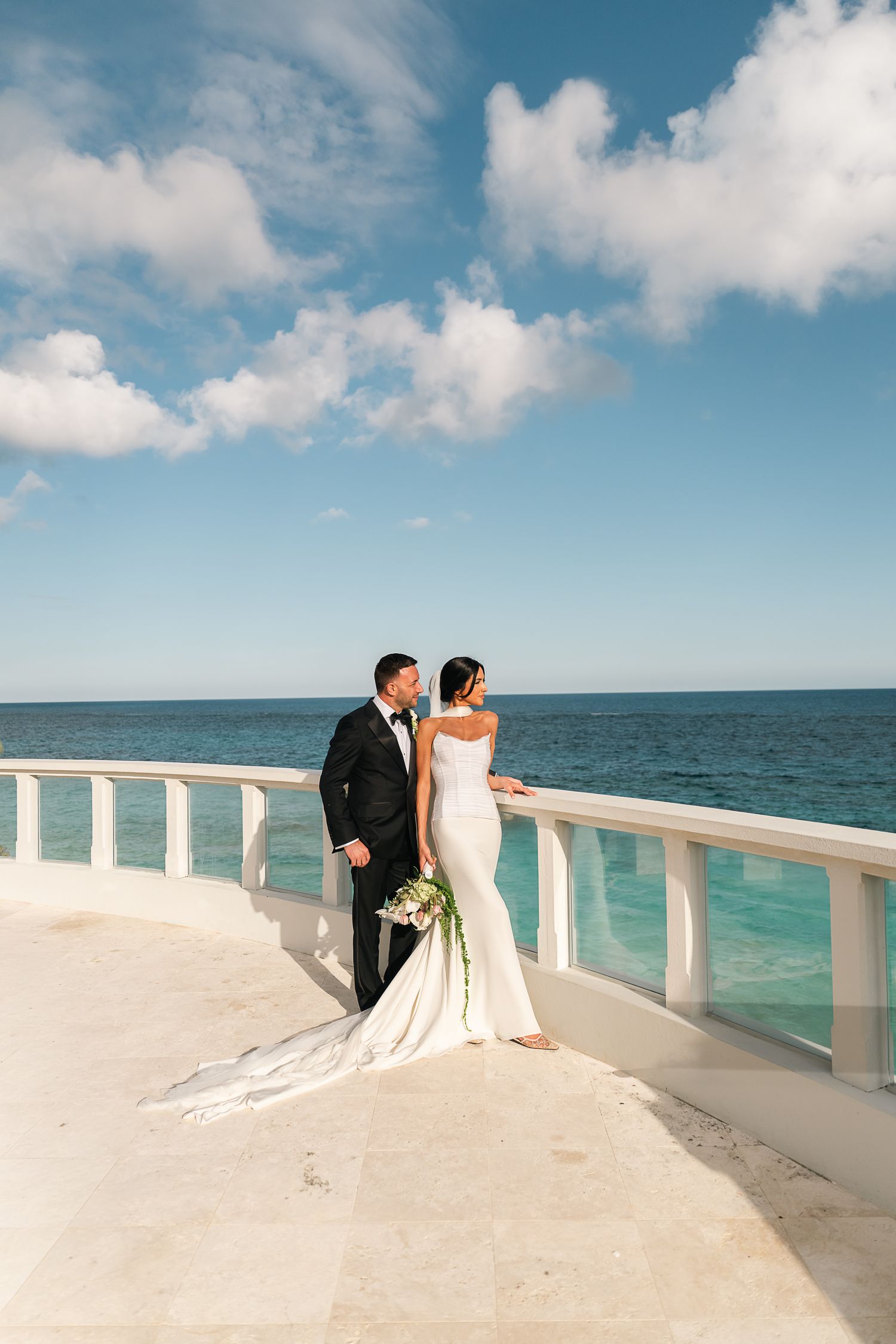 Bride and groom standing on elegant oceanfront terrace, bride holding bouquet with turquoise sea and blue sky backdrop.