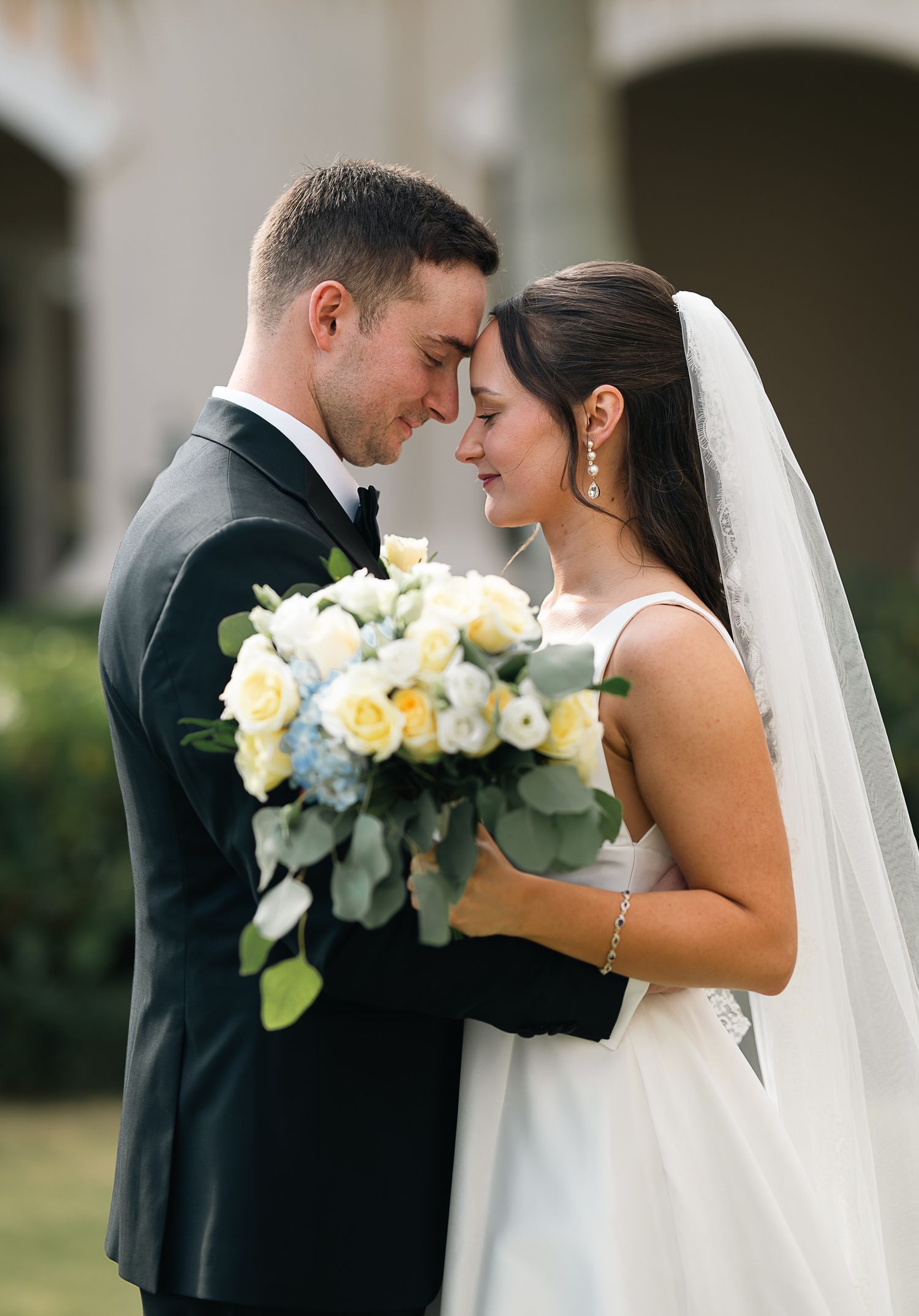 Bride and groom touch foreheads tenderly while holding yellow and white floral bouquet.